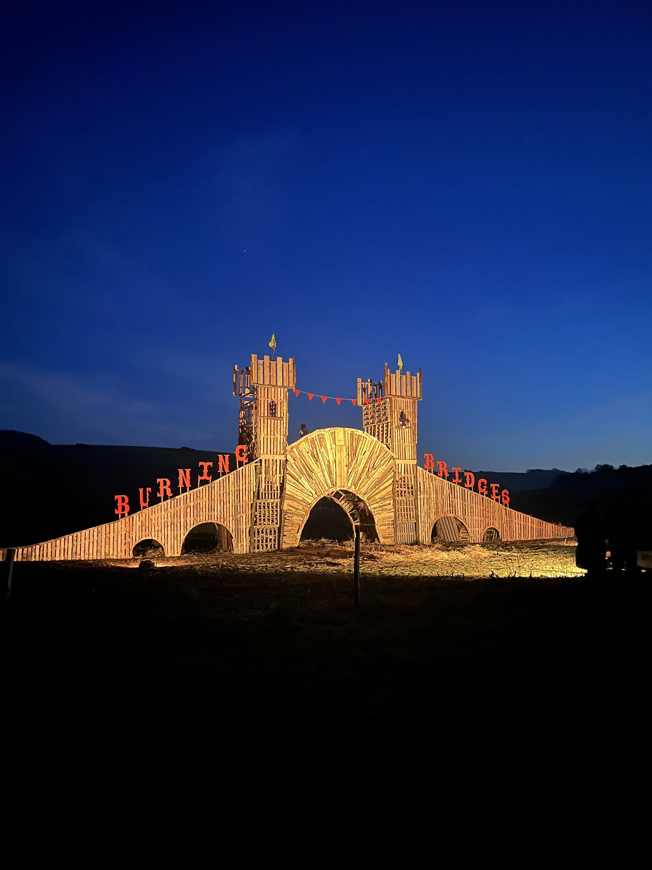 Wooden castle-like structure decorated with red letters spelling 'BURNEING BRIDGES' and illuminated at night against a dark sky.