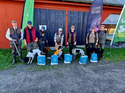Group of people with dogs and blue gift bags outdoors in front of a barn and colorful flags.