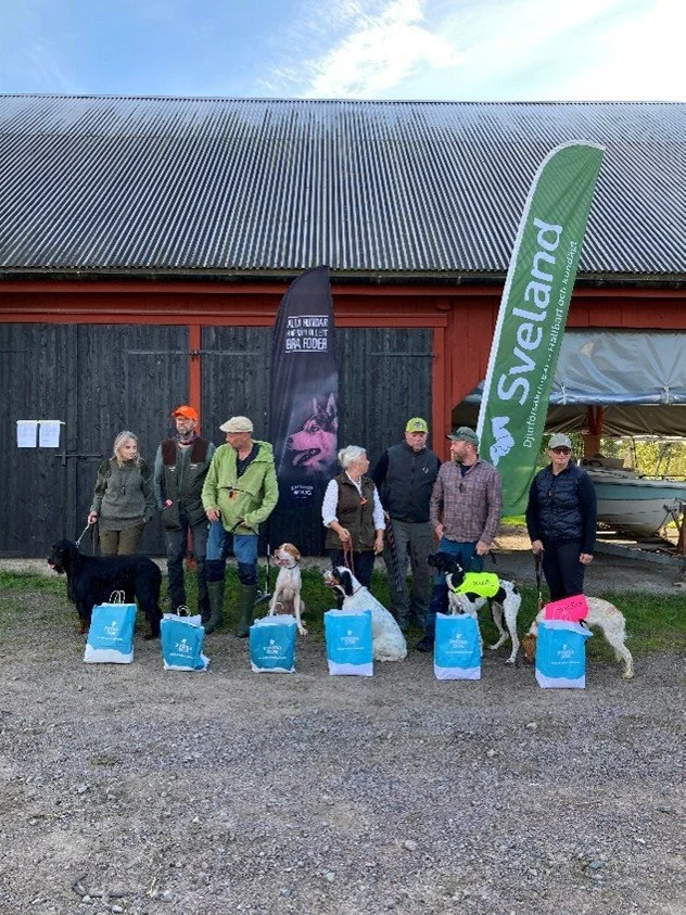 Group of people with dogs and prize bags, standing outdoors in front of a barn with flags that say 'Svealand' and pet-themed text.
