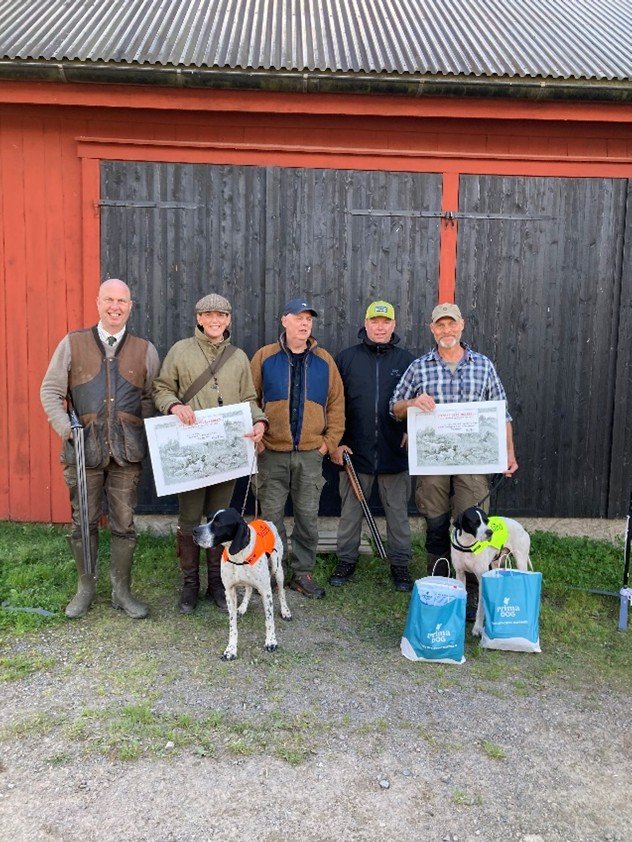 Five people standing outdoors in front of a large black and red wooden barn door. The person second from the left and the person on the far right are holding framed pictures. They are accompanied by two dogs on leashes, with one dog wearing an orange vest and the other a yellow vest. Two blue bags are on the ground in front of the dogs.