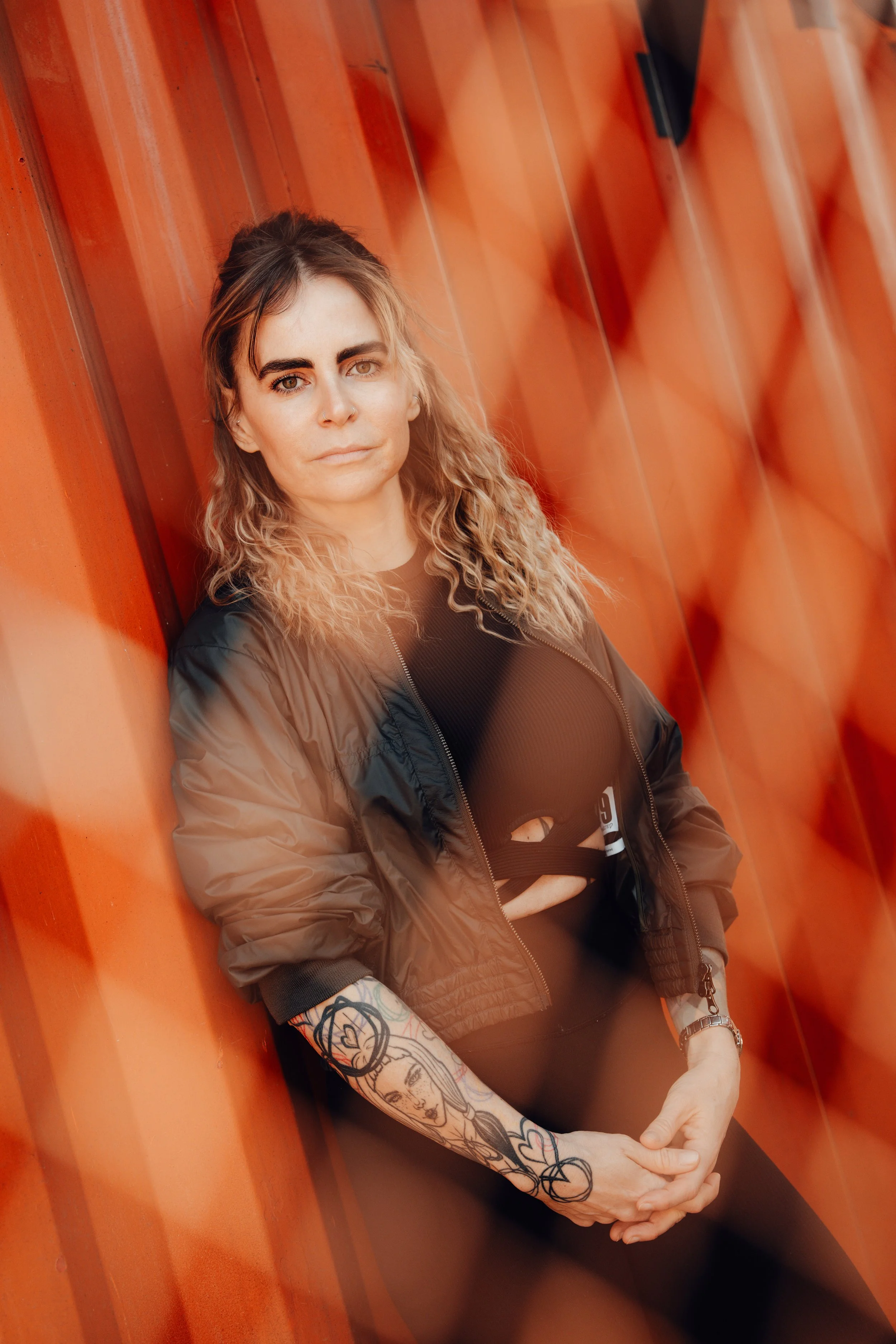 Junge Frau mit Tattoo am Arm, schwarze Haare, trägt eine schwarze Spitzenkleidung und eine Bomberjacke, sitzt an einer roten Wand und schaut in die Kamera.