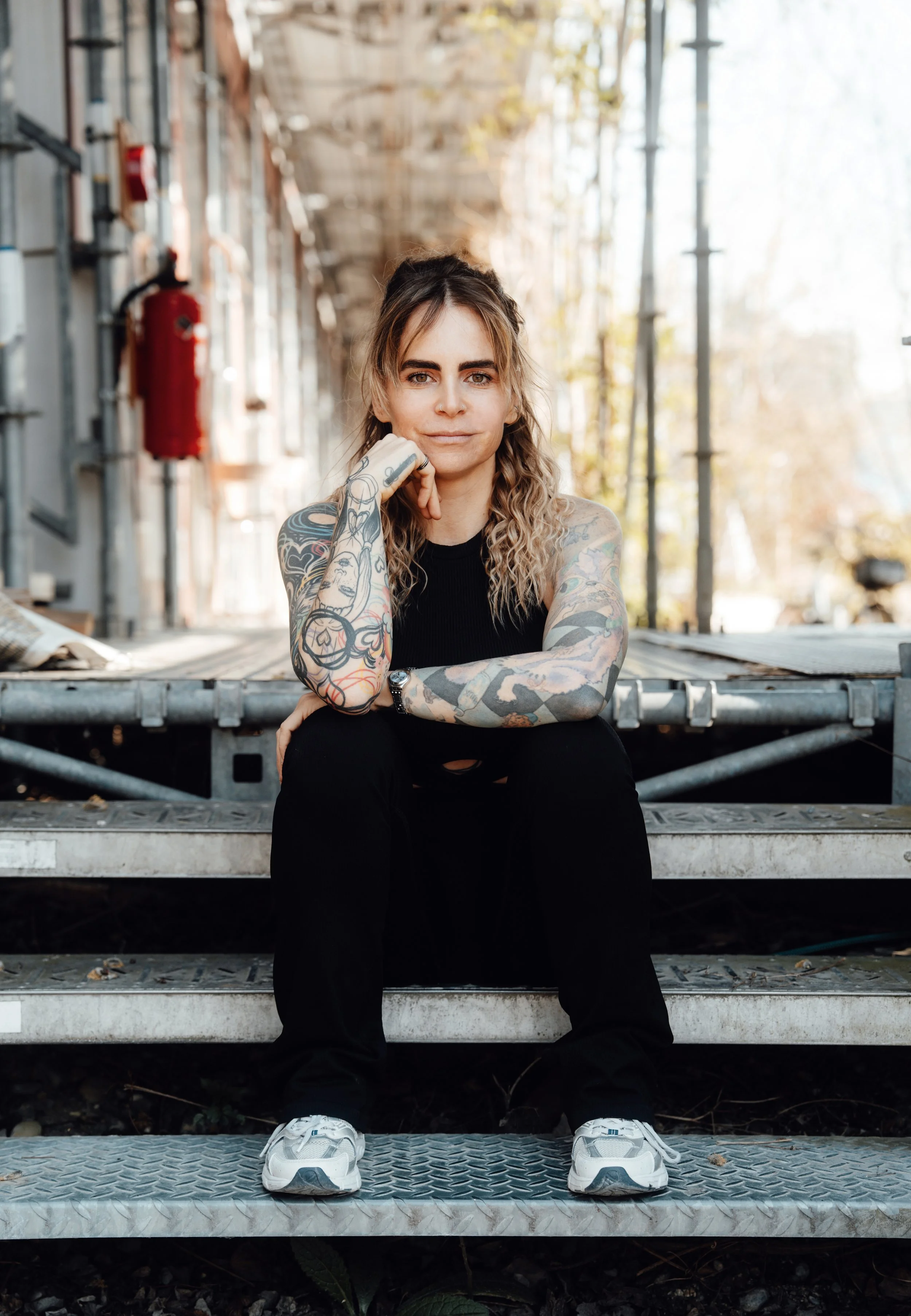 Junge Frau mit tattoos sitzend auf Metalltreppe im Freien, lächelnd, mit Baum und Gebäude im Hintergrund.