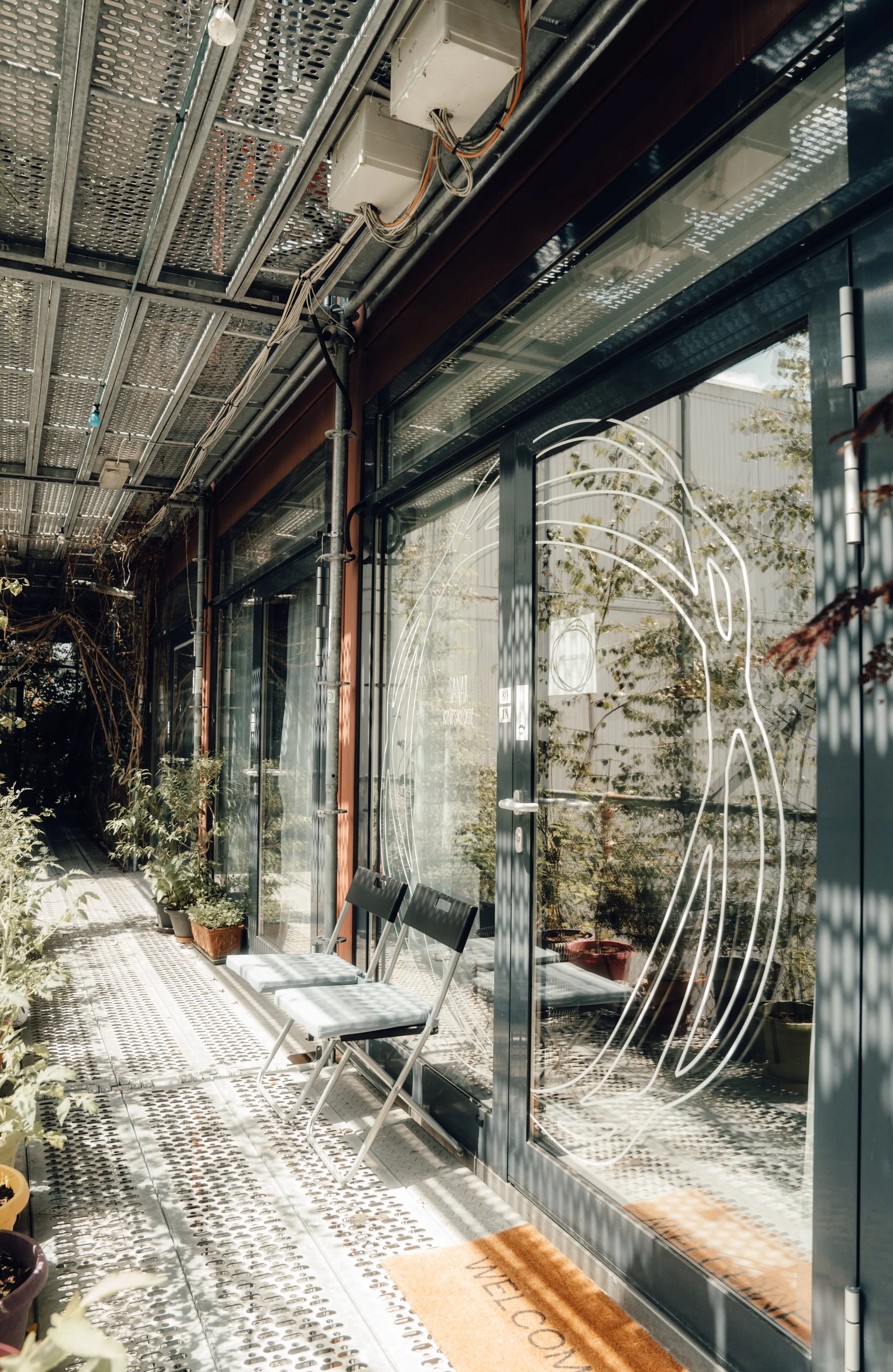 Blick auf einen begrünten Außenbereich eines Cafés mit großen Glasfenstern, in denen ein Kaffee-Logo zu sehen ist. Es stehen zwei schwarze Klappstühle vor den Fenstern, umgeben von Pflanzen in Töpfen. Das Design ist modern und freundlich, mit einem Eingangsmatte, auf der "Willkommen" steht.