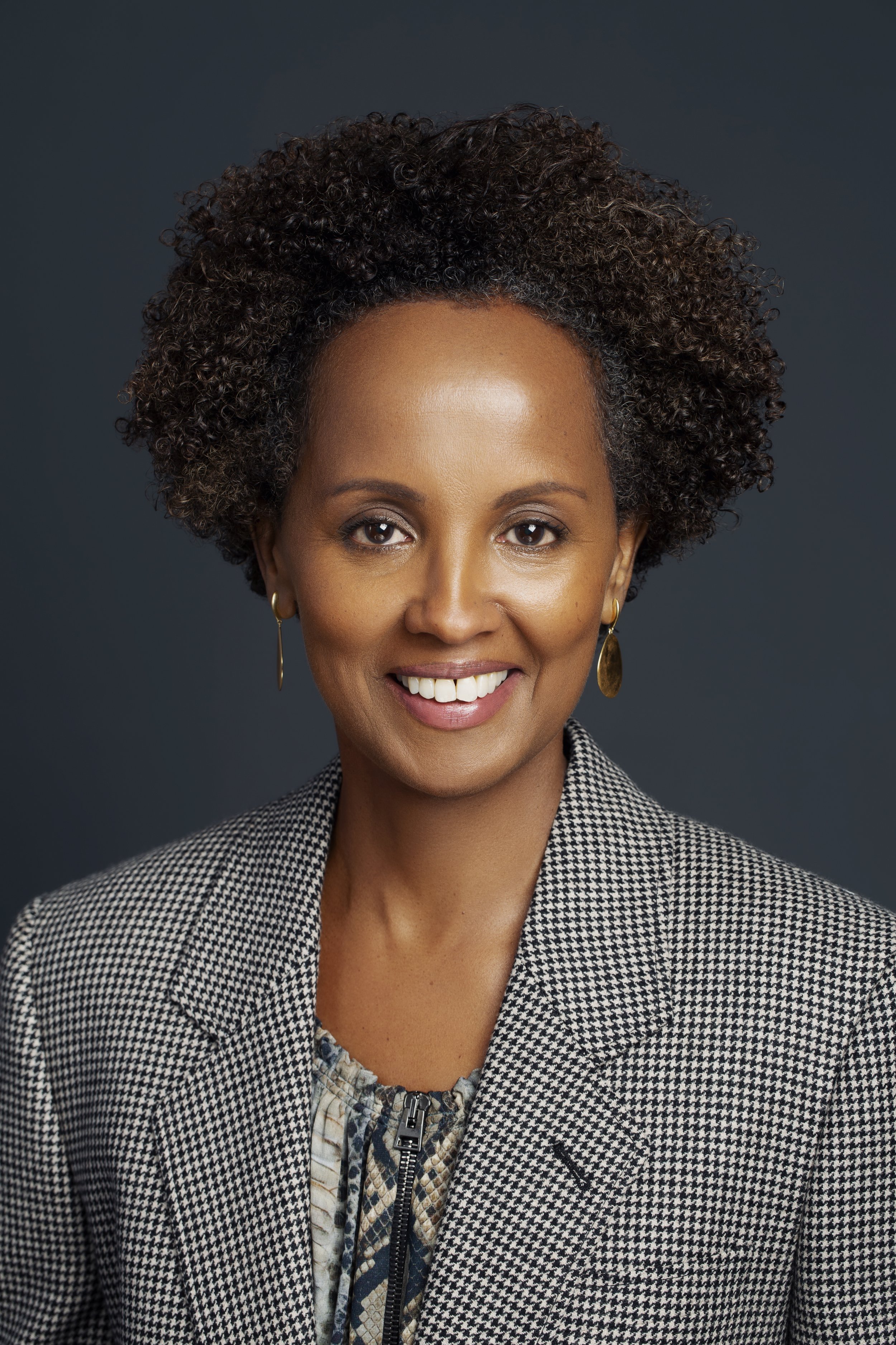 A woman with dark skin and curly hair smiling at the camera, wearing gold earrings and a houndstooth blazer over a patterned top, set against a dark background.