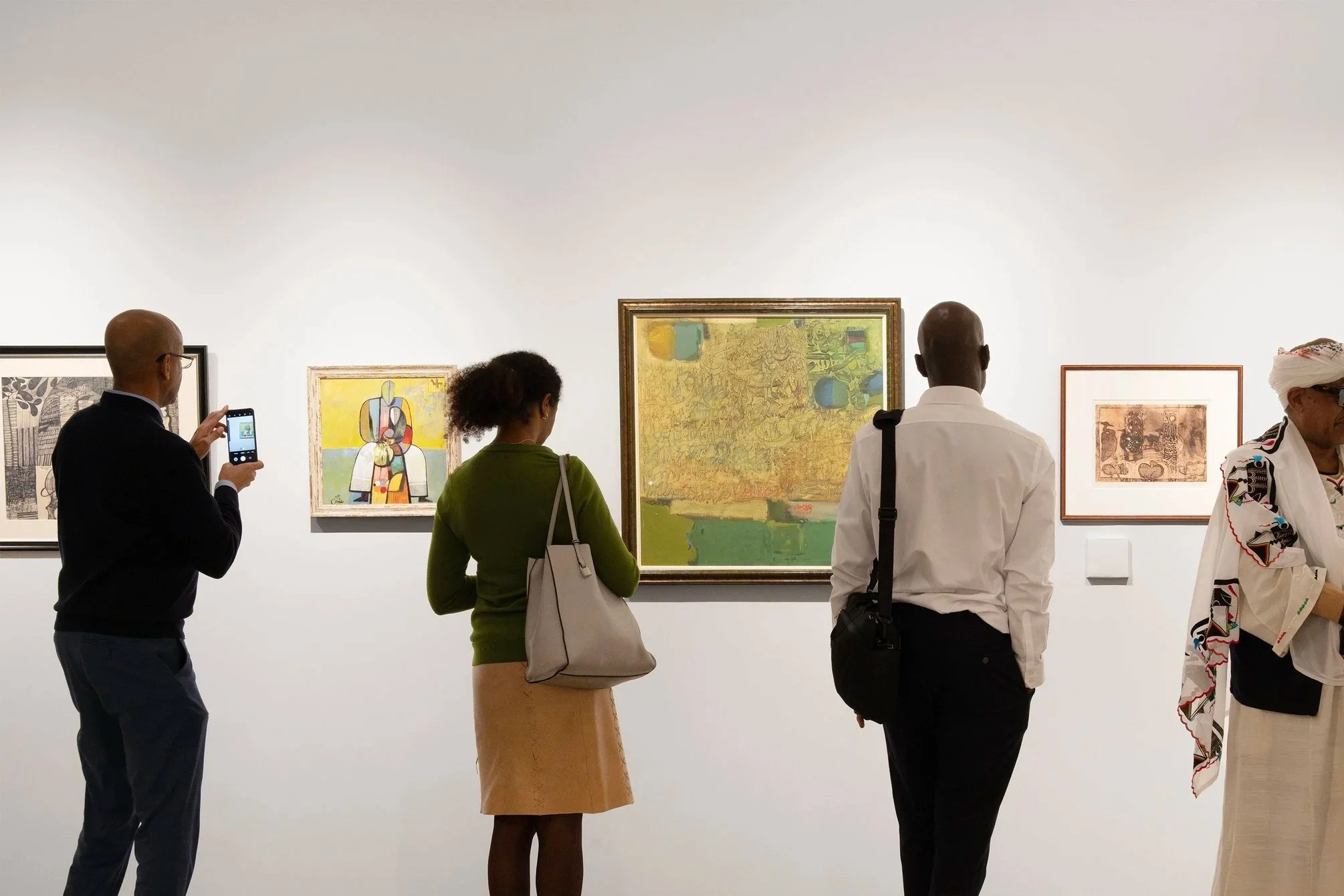 People viewing artwork in an art gallery, with one person taking a photo of a picture on the wall.