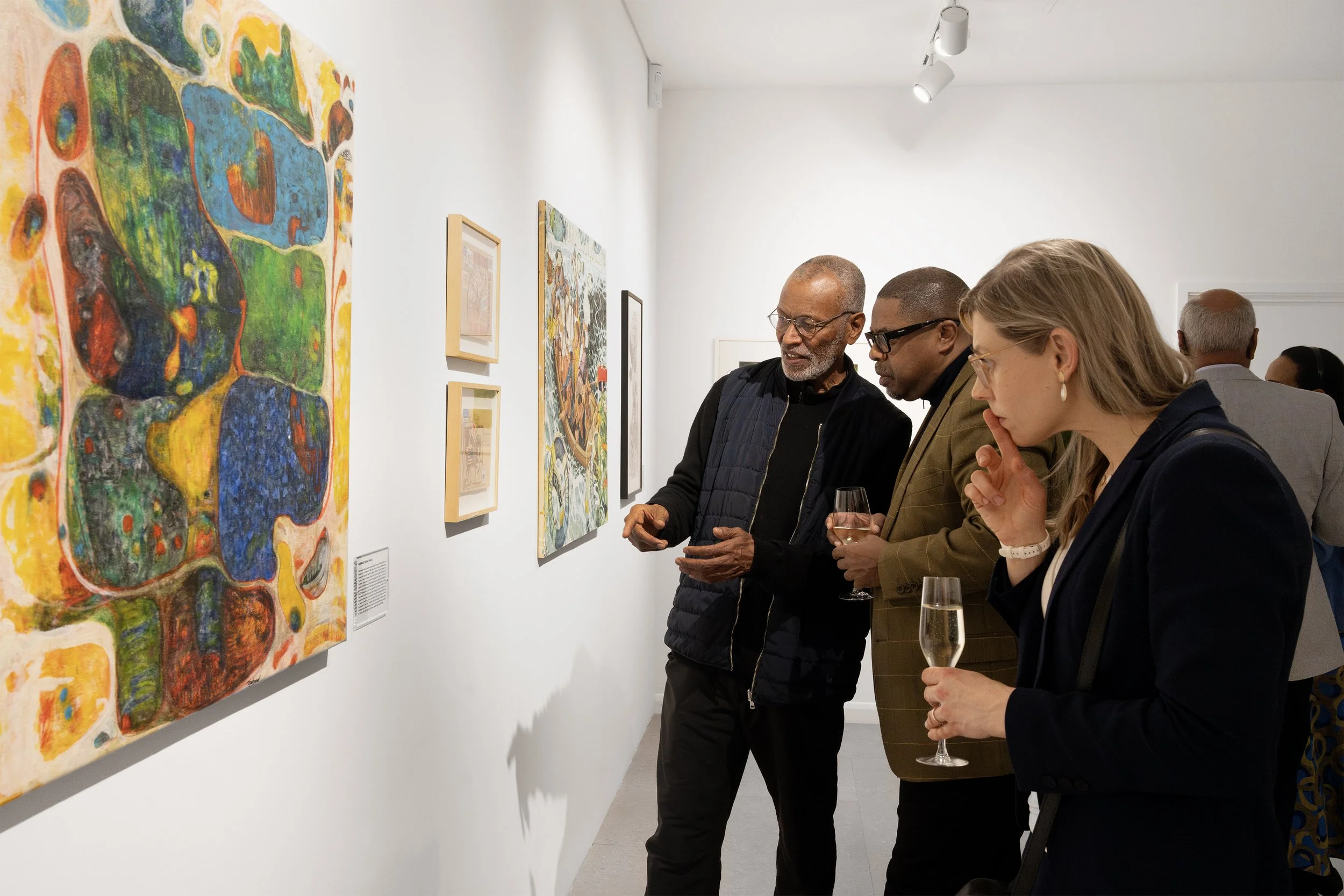A group of people at an art gallery viewing colorful paintings on the wall. They are holding glasses of champagne and appear engaged in conversation.