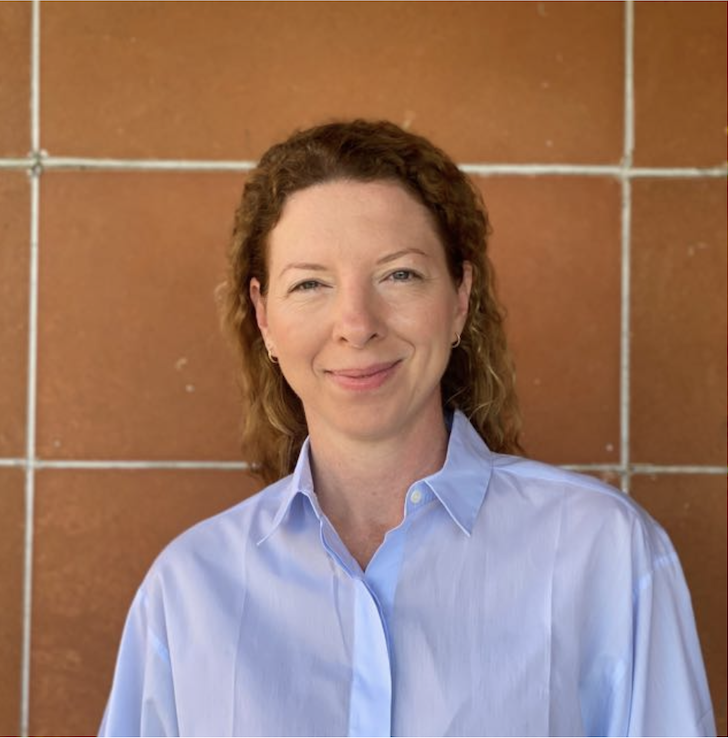 A woman with curly red hair smiling in front of a brown tiled wall, wearing a light blue button-up shirt.