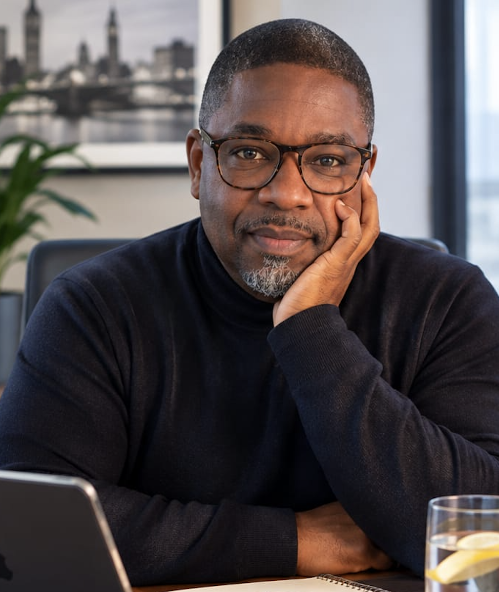 A man with glasses and a short beard resting his face on his hand, seated at a desk in an office with a cityscape picture on the wall behind him and a glass of water with lemon in the foreground.