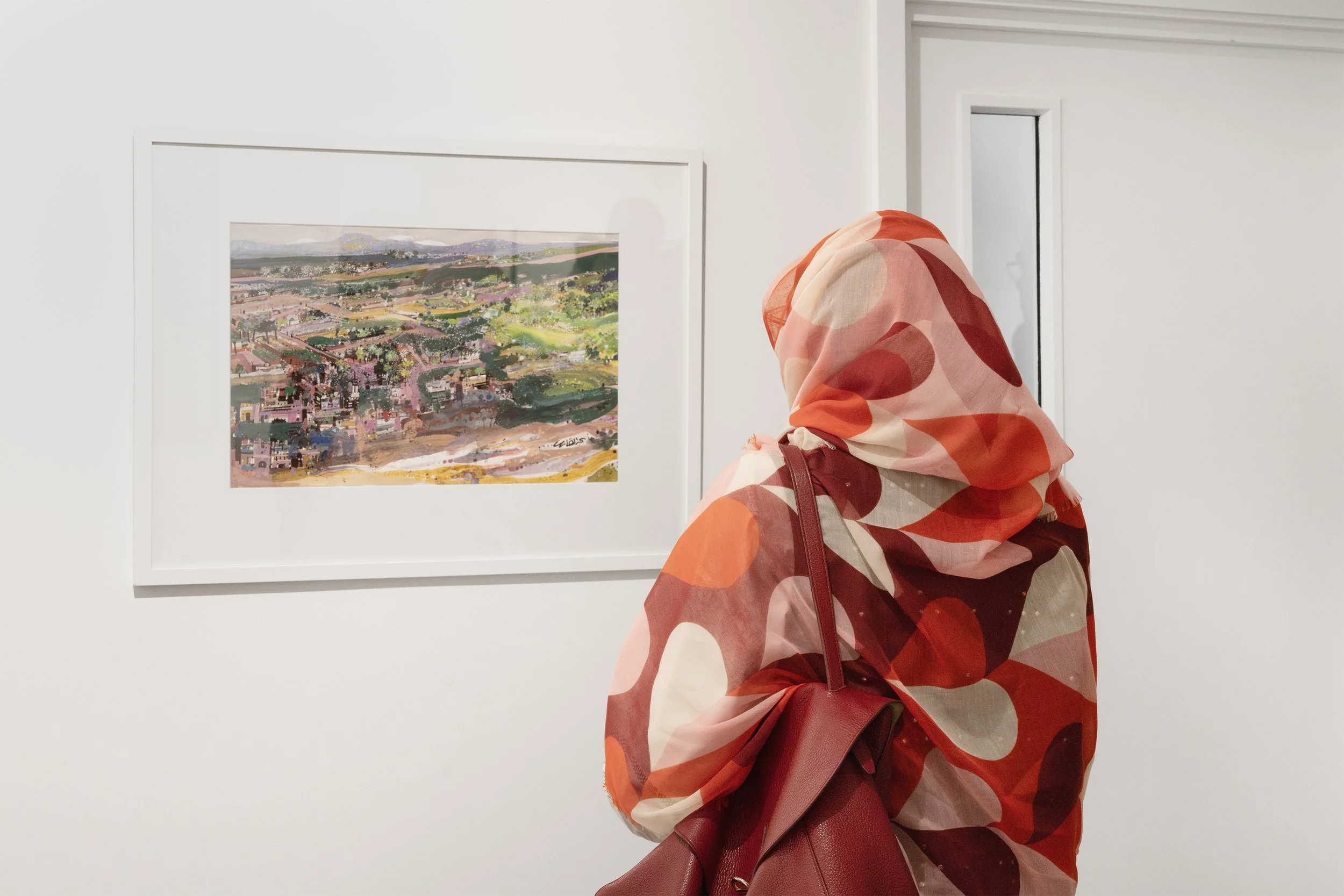 A person wearing a colorful, patterned hijab and carrying a red handbag, viewing a colorful landscape painting in an art gallery.