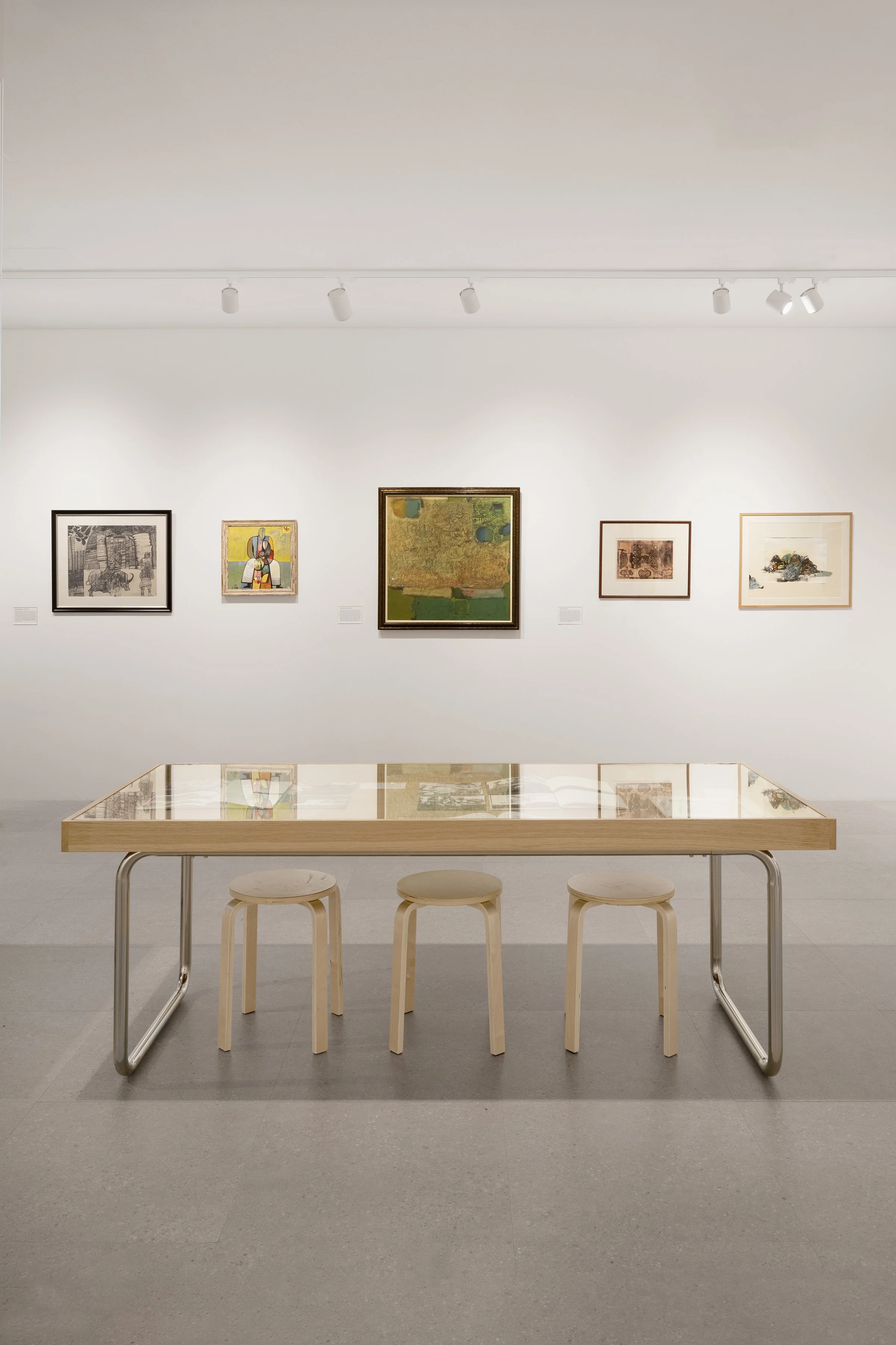 An art gallery with five paintings on a white wall and a glass-topped table with three stools in the foreground.