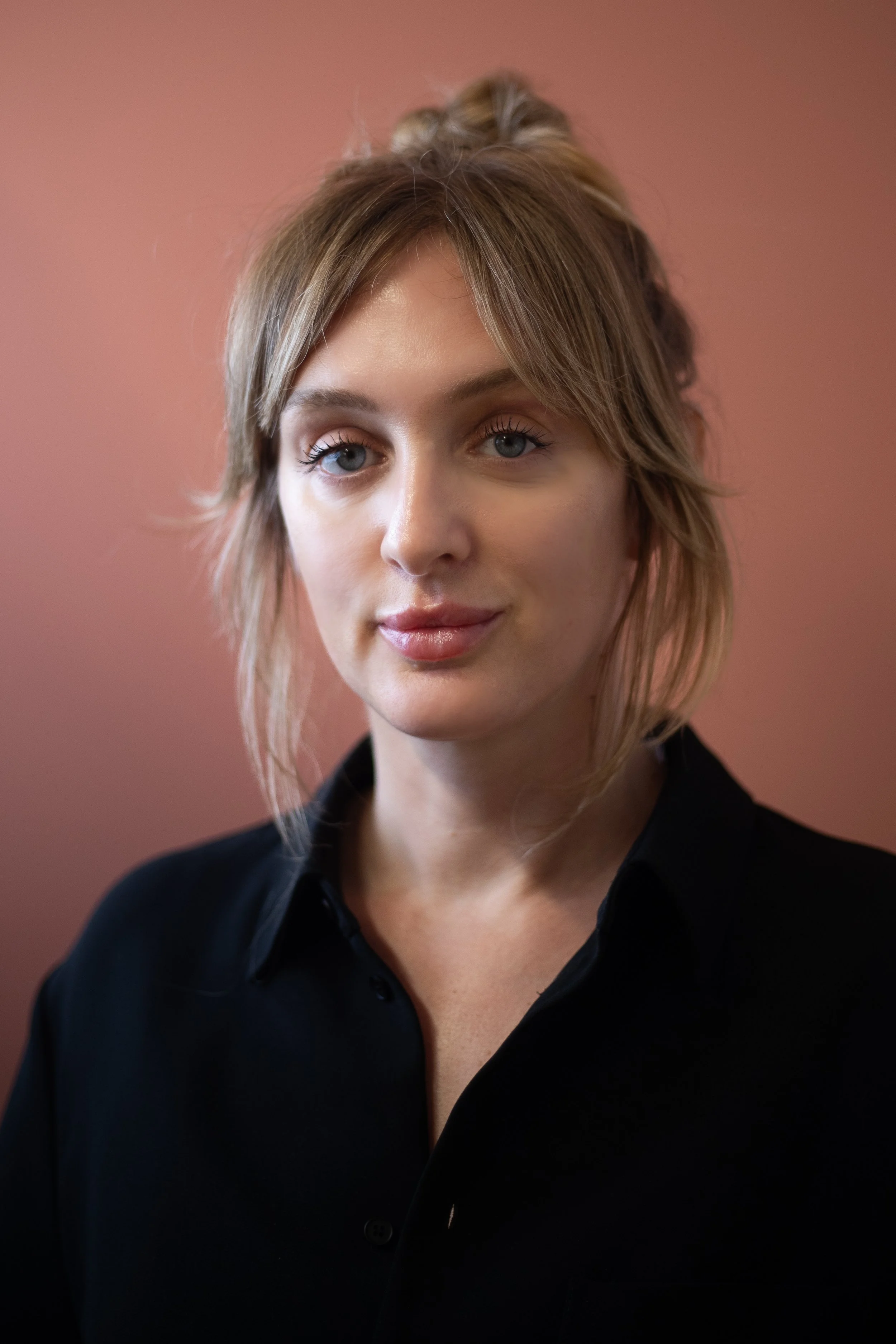 A woman with blonde hair styled in a loose updo, wearing a black shirt, standing against a pink background.