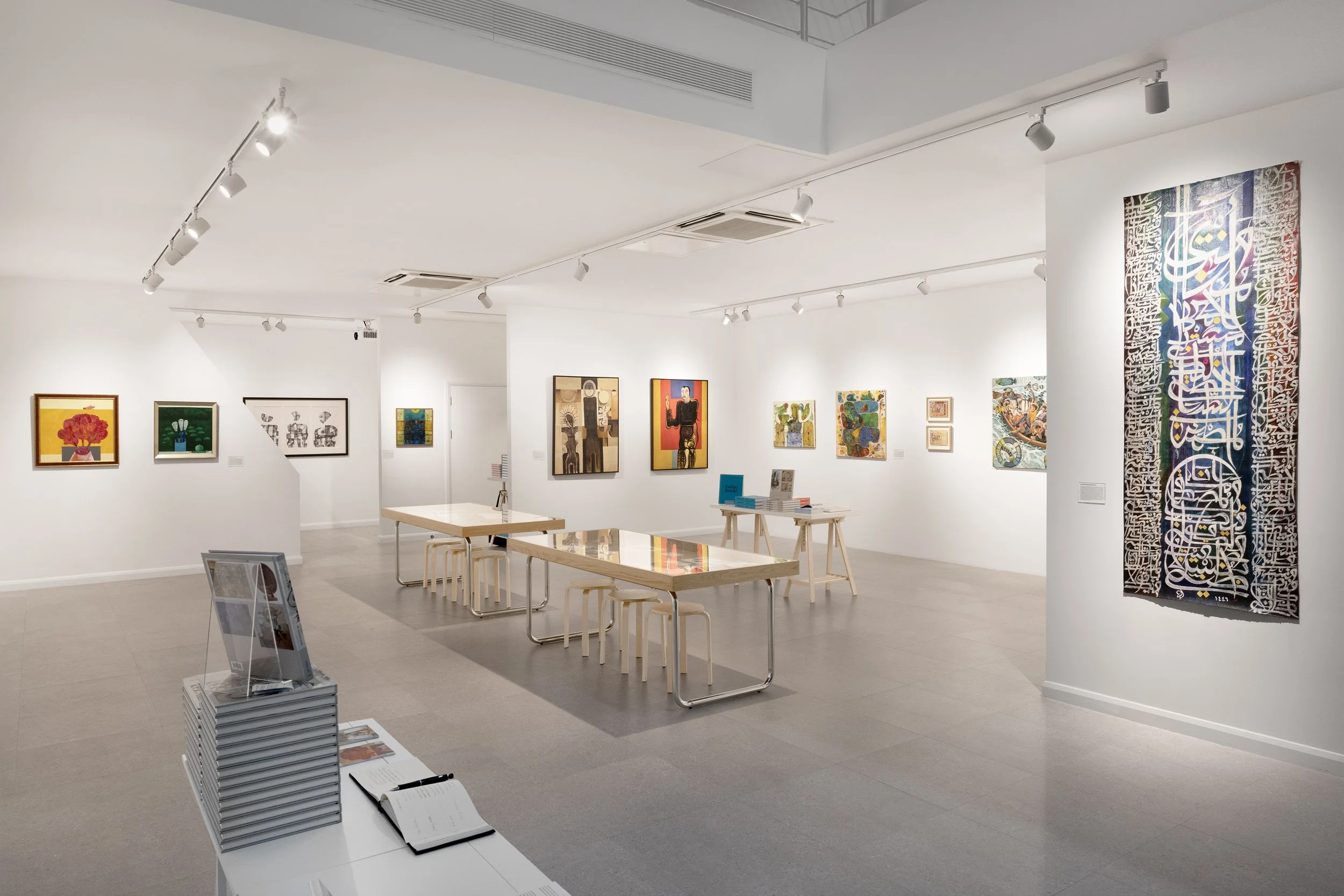 Empty art gallery with white walls displaying colorful paintings and calligraphy art. There are tables with books and a display stand with brochures, all in a bright, modern setting.