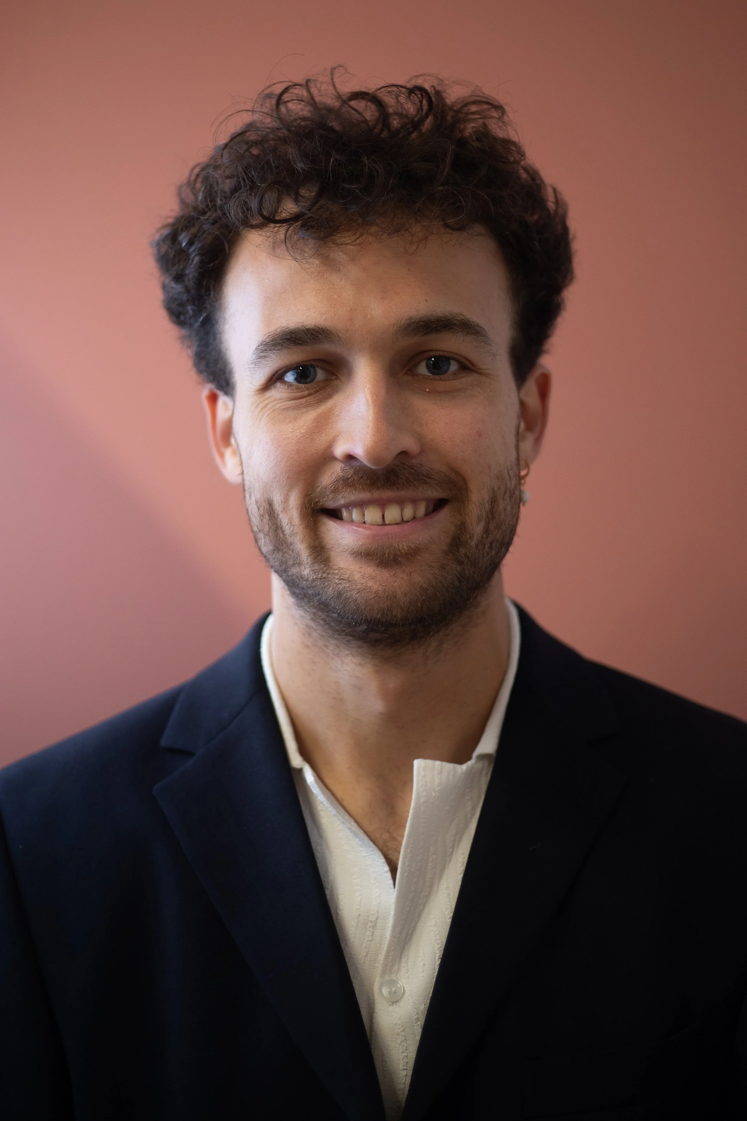 A young man with curly hair, light skin, blue eyes, and a beard, smiling in a formal black suit and white shirt, against a soft pinkish background.