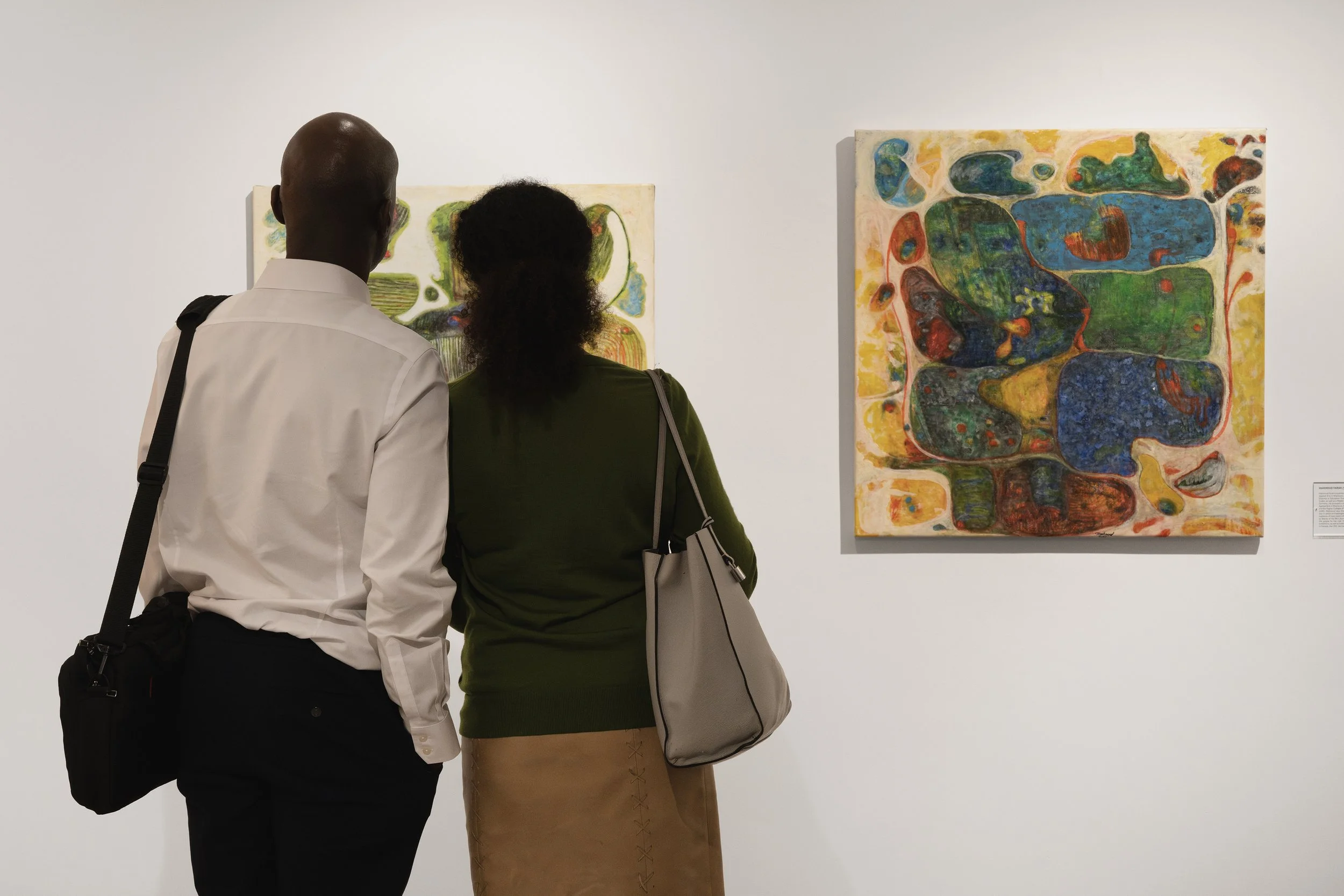 Two people standing next to each other in a gallery looking at paintings.