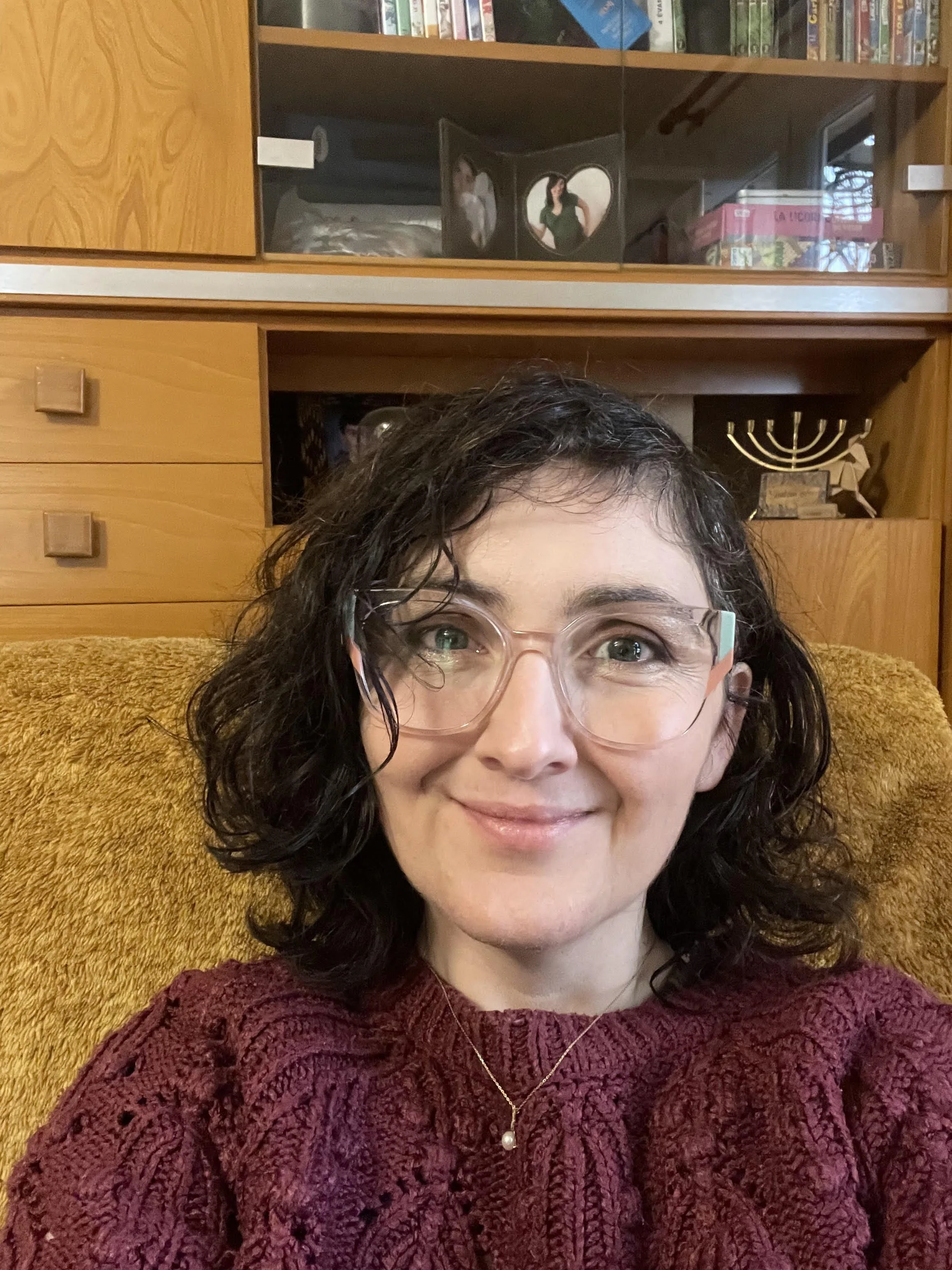 Femme souriante portant des lunettes, portant un pull en tricot rouge, assise dans un fauteuil en tissu orangé avec une étagère en bois en arrière-plan.