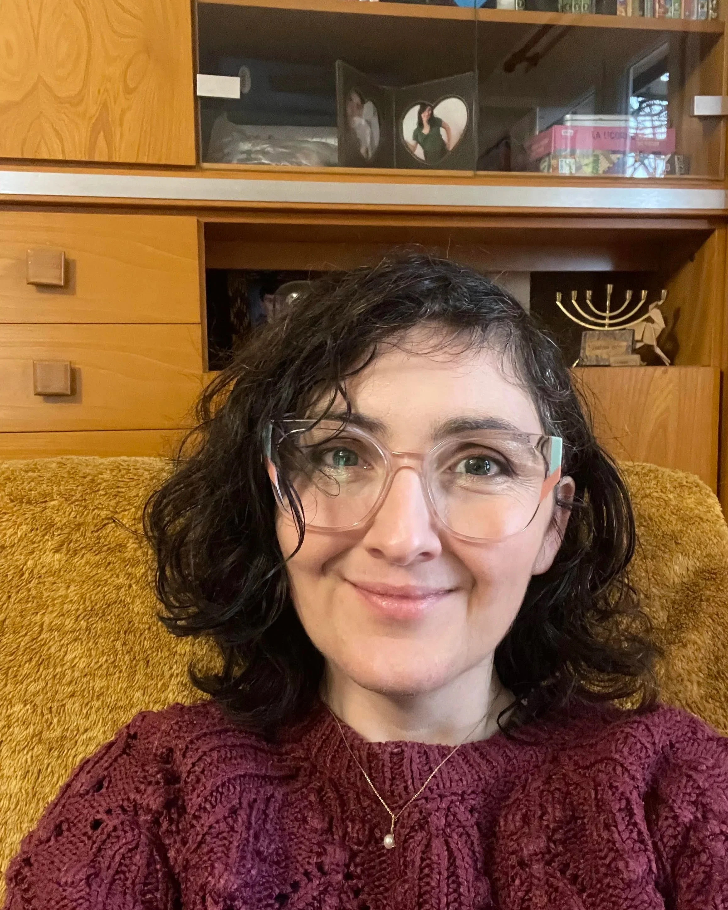 Une femme souriante avec des lunettes, portant un pull bordeaux, assise sur un canapé jaune, avec une bibliothèque en bois en arrière-plan.