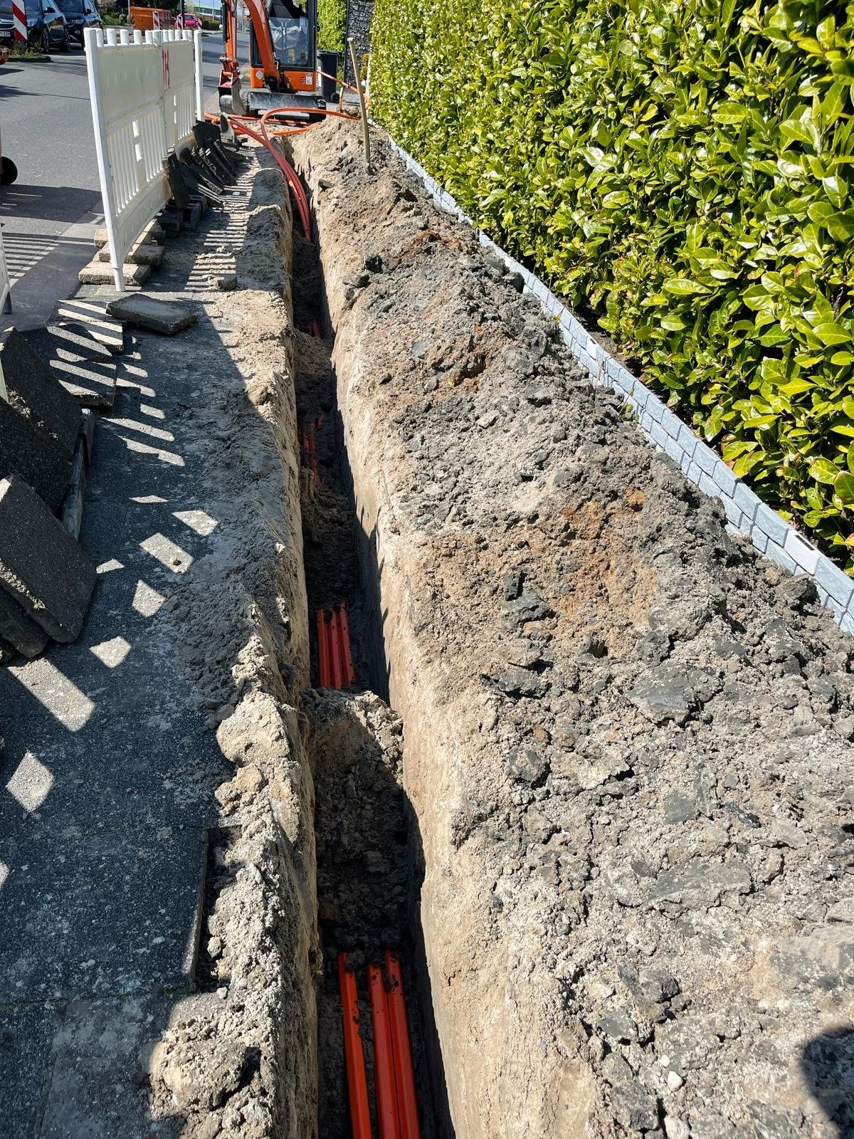 Construction workers are installing underground utility lines; a trench is dug along a sidewalk with orange conduit pipes inside.