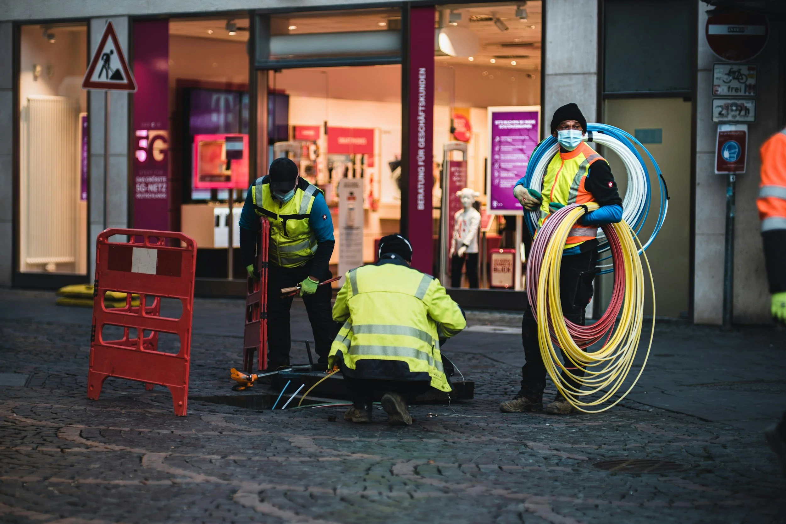 Construction workers in high-visibility jackets and masks working on underground electrical or communication cables in a city street, with a shop in the background.
