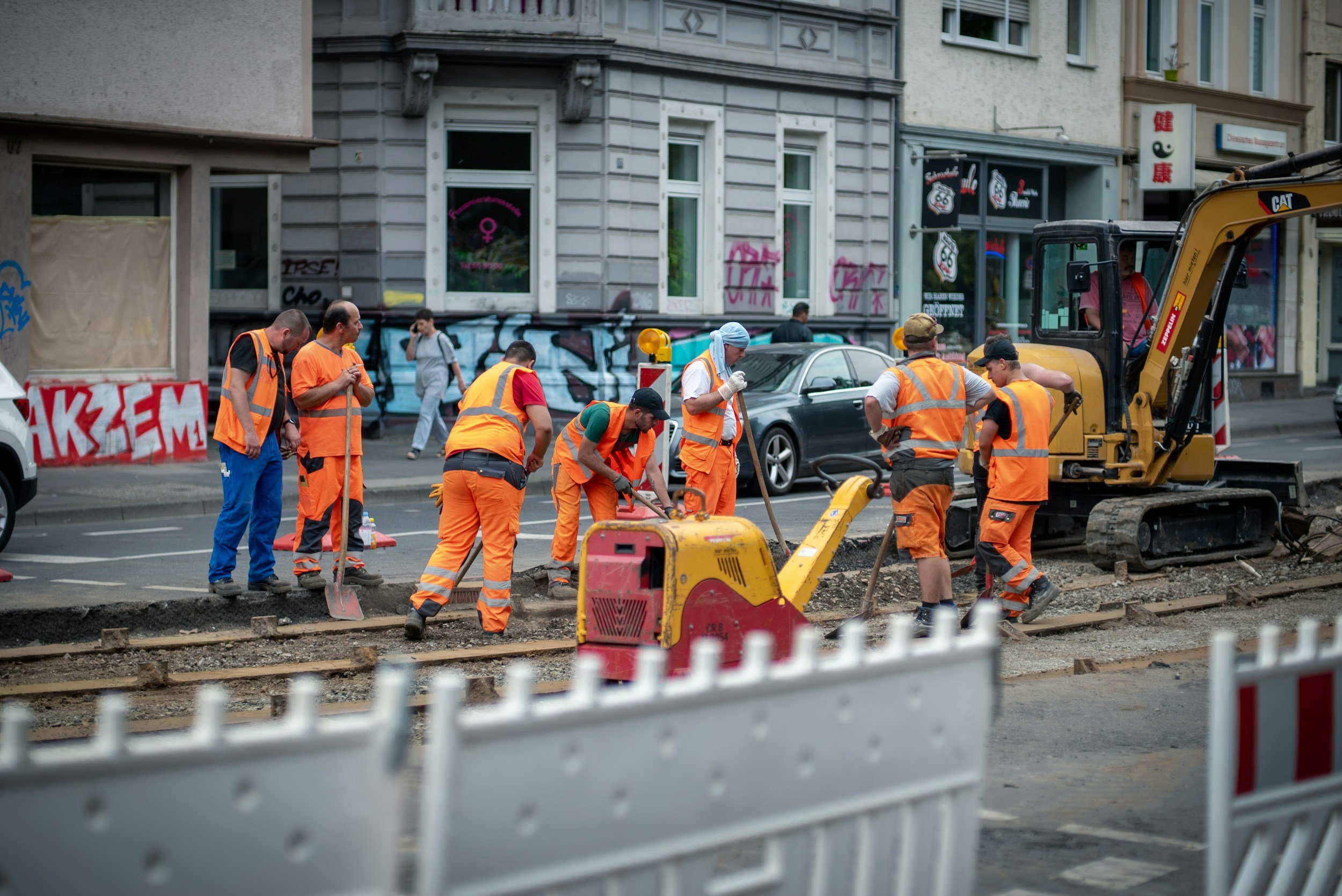 Construction workers in orange safety vests working on street rail line, with a small construction vehicle and parked cars in the background.