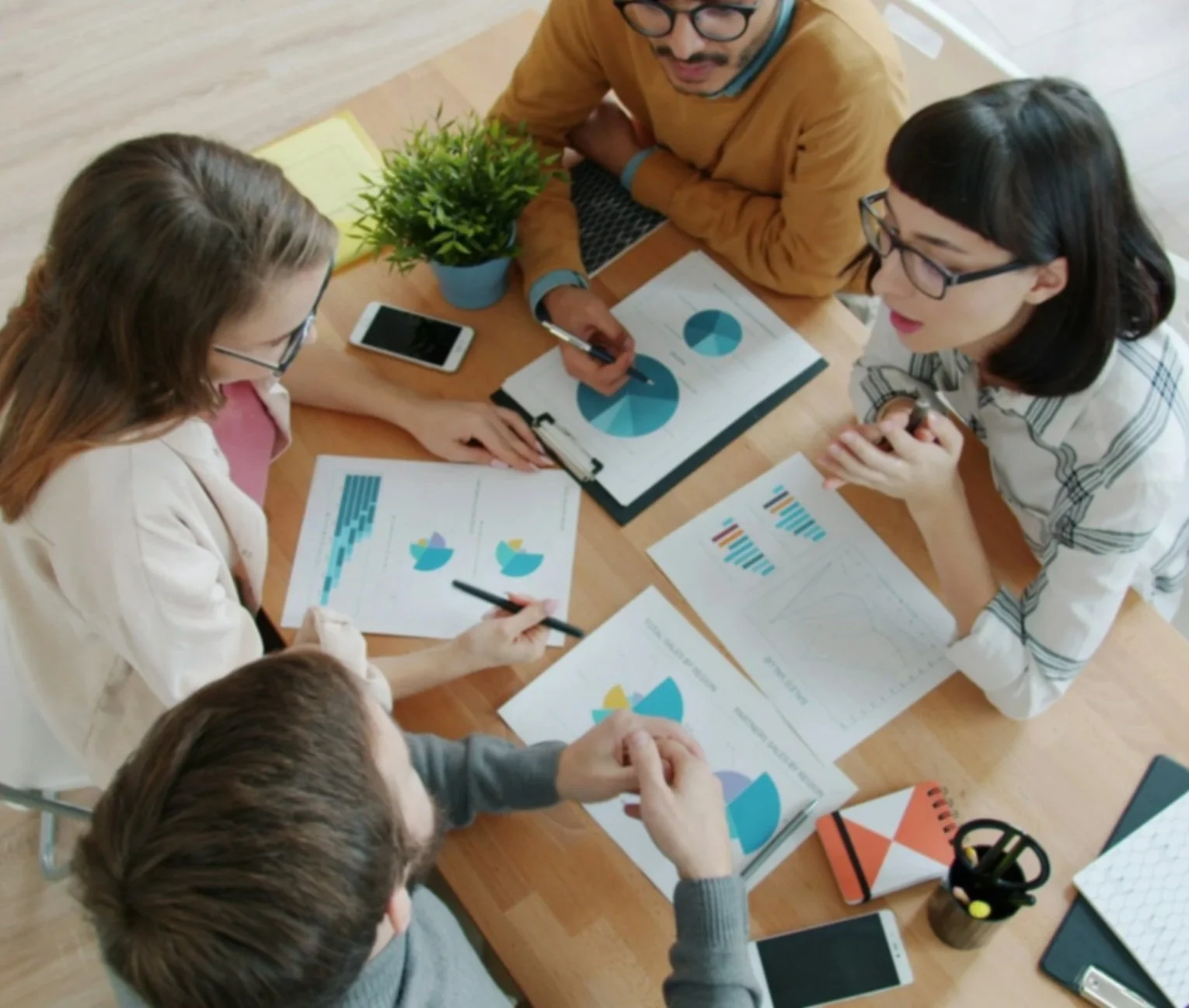 Five people sitting around a wooden table engaged in a business meeting, discussing charts and graphs with printed data, tablets, and a potted plant on the table.