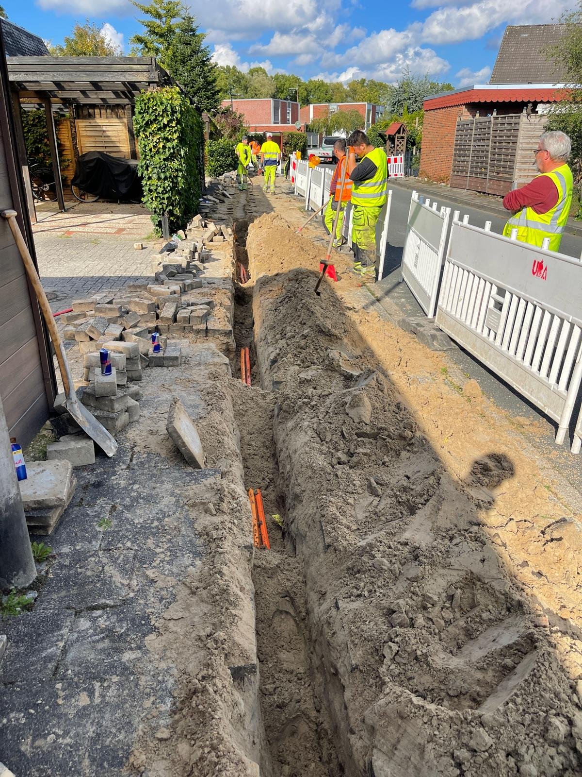Construction workers digging a trench on a residential street for underground utility work, with equipment and materials nearby, and barriers blocking part of the road.