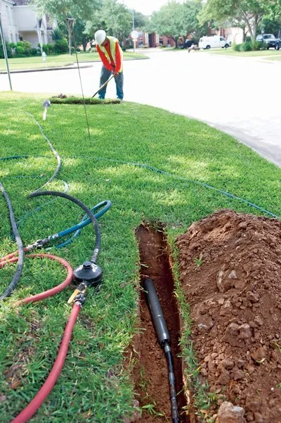 A worker in safety gear digging a trench on a grassy area with equipment and hoses nearby, likely for underground cable work.