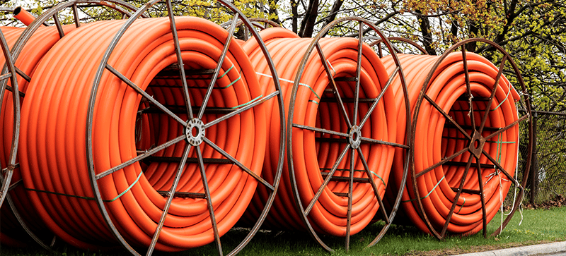 Large rolls of orange electrical conduit stored on metal spools outdoors on grass, with trees and a fence in the background.