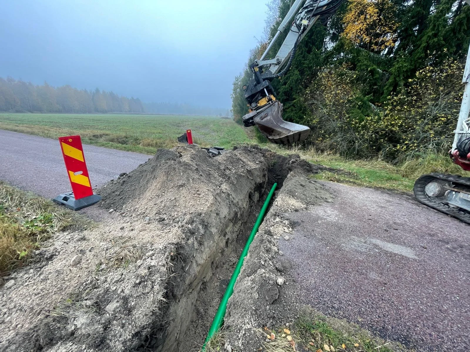 Construction site on a rural road with an open trench where a green pipe is being laid, a small excavator, and safety barriers.
