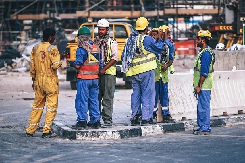 Group of construction workers gathered on a road construction site, wearing safety vests and hard hats, with construction equipment and a building structure in the background.