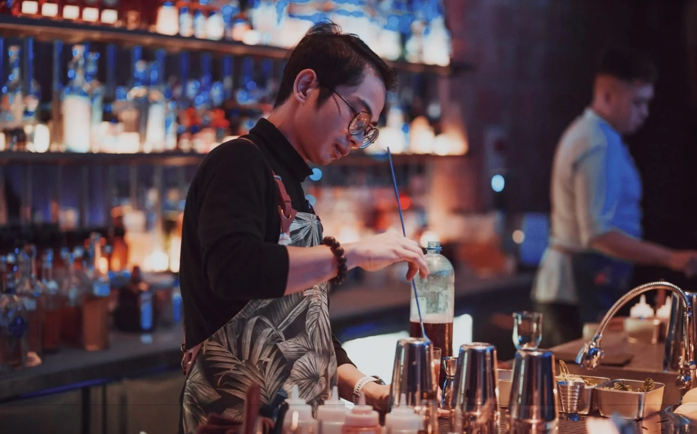 A bartender mixing a drink at a bar with bottles on shelves behind him and another person in the background.