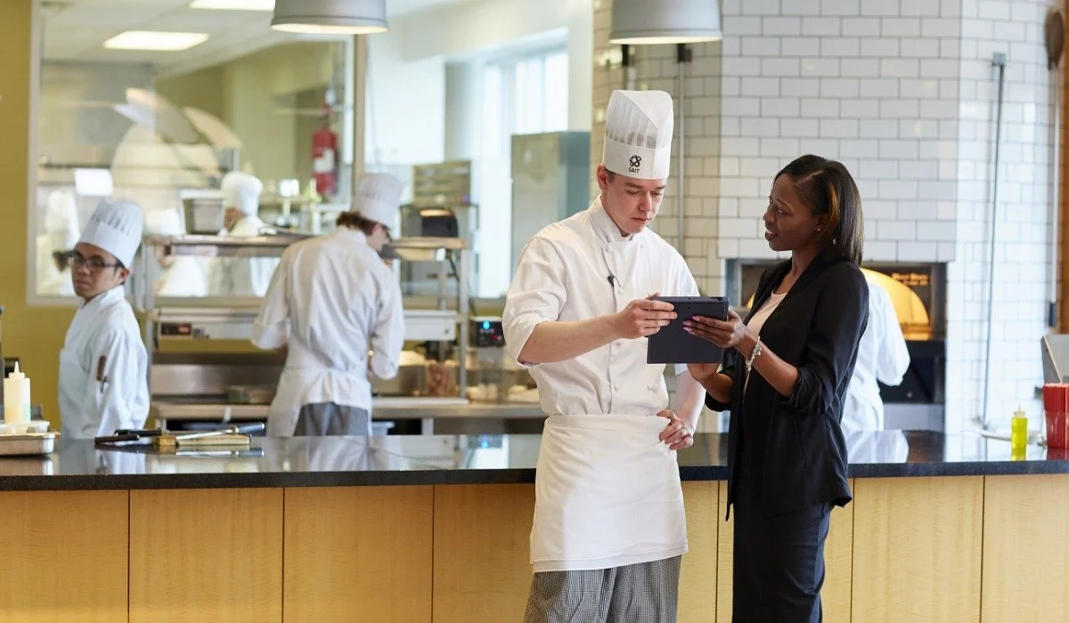 A chef in a white uniform and hat showing something on a tablet to a woman in business attire in a restaurant kitchen with other chefs working in the background.