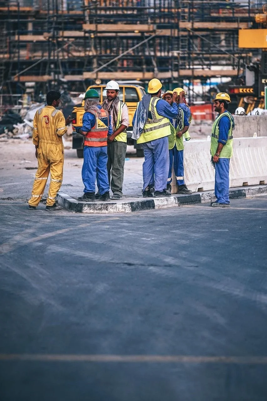 Group of construction workers gathered on a road construction site, wearing safety vests and hard hats, with construction equipment and a building structure in the background.