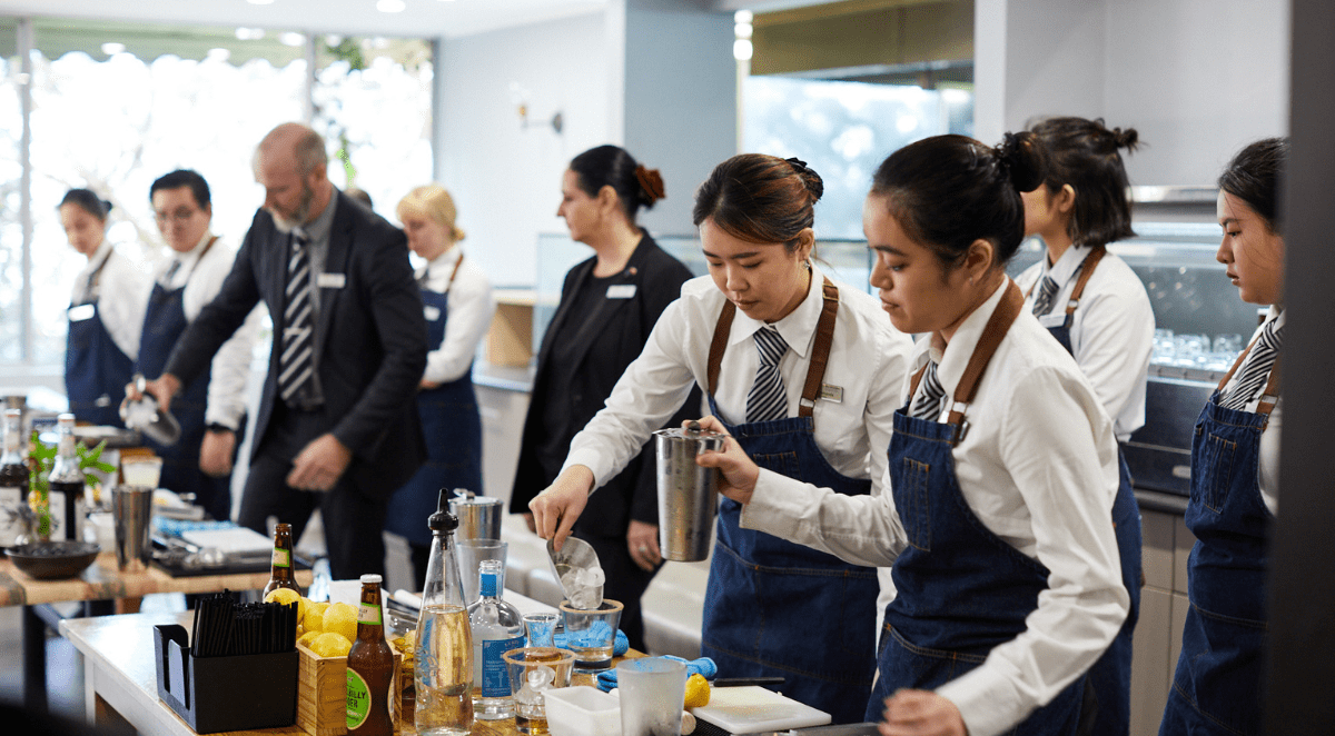 A group of people in formal attire, wearing blue aprons, participating in a cooking class or demonstration in a modern kitchen with large windows.