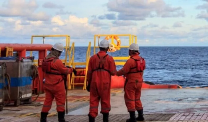 Three workers in red coveralls and white helmets standing on a ship's deck near the ocean, engaged in a discussion.