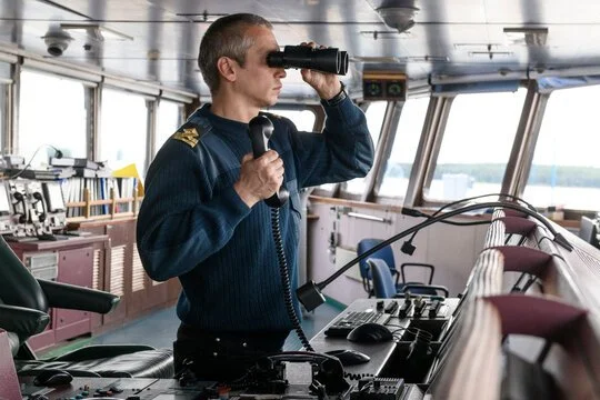 A man in a uniform stands in a ship's control room, looking through binoculars.