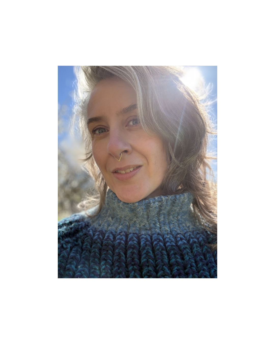 Close-up of a woman smiling outdoors, with sunlight shining behind her, wearing a knitted sweater and a septum piercing.
