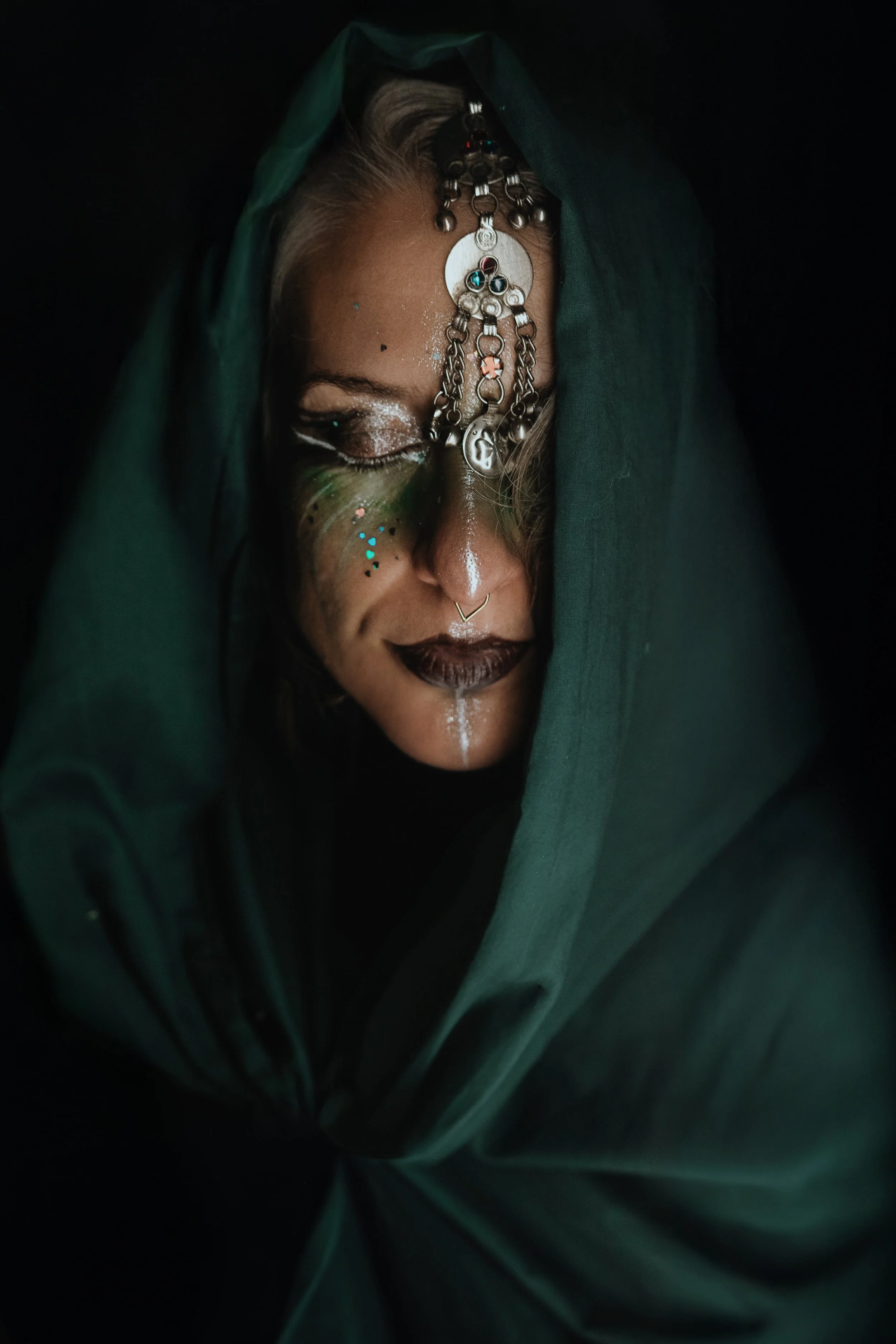 A person with a metallic headpiece wearing dark makeup and glitter, partly covered by a dark green cloth, with a dark background.