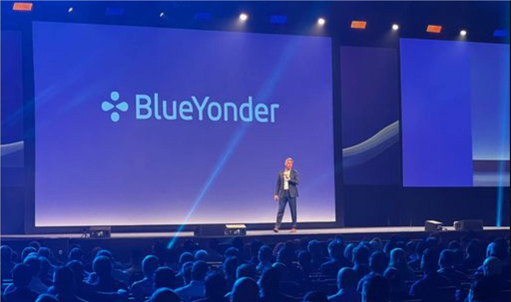 A speaker presenting on a large stage at a conference, with a screen displaying the BlueYonder logo behind him, in front of an audience.