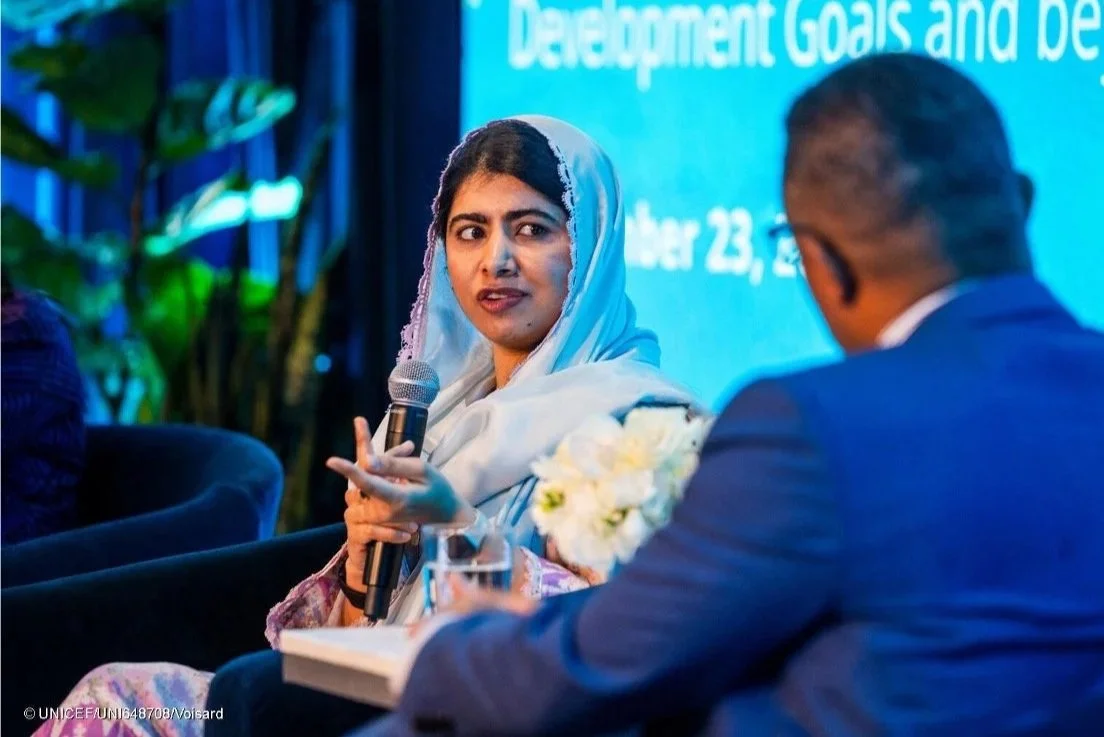 A woman wearing a grey headscarf is speaking into a microphone during a panel discussion, while a man in a suit listens. A blue screen with text is in the background.