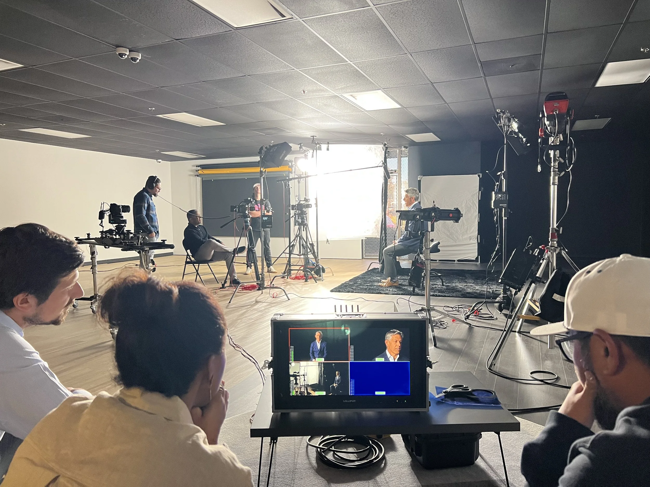 A behind-the-scenes view of a video recording studio with a seated man being filmed, crew members operating cameras, and an overall setup for a video shoot or interview.