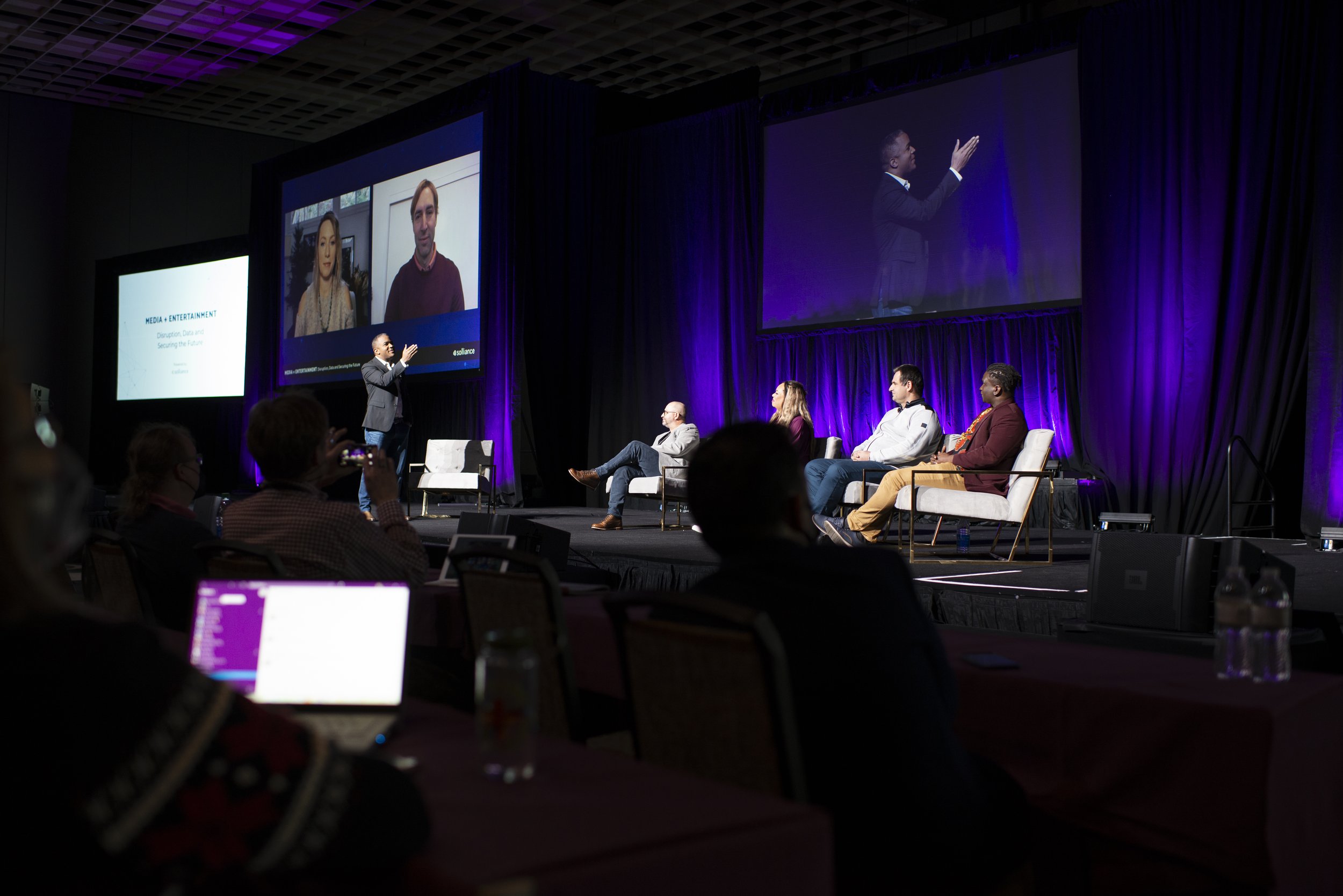 A panel discussion at a conference featuring five speakers on stage, one of whom is standing and speaking into a microphone while the others sit in chairs. Large screens behind display video feeds and presentation slides, with purple and black curtains as the backdrop.