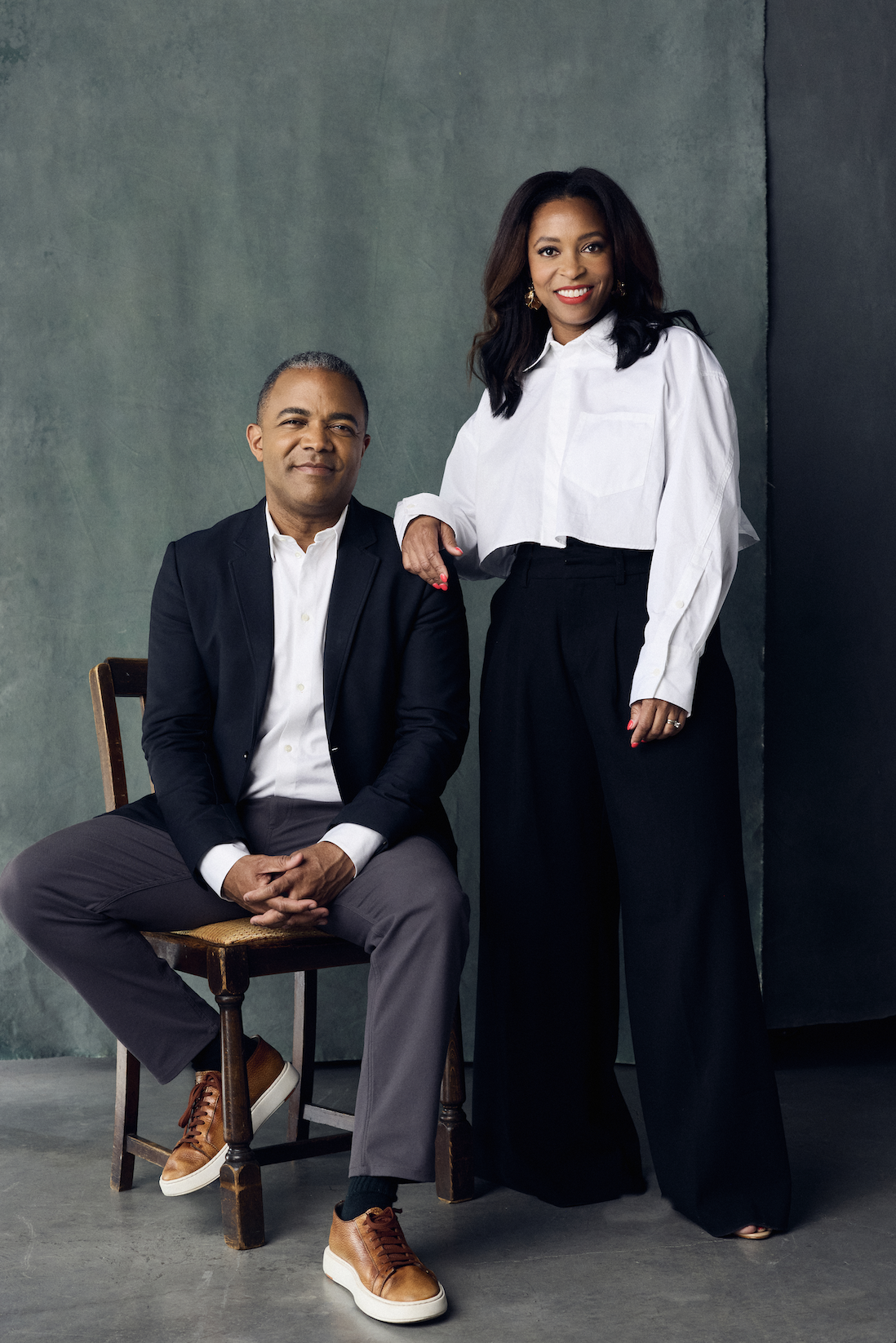 A professional man in a dark blazer and gray pants seated on a wooden chair, and a woman in a white blouse and black wide-leg pants standing beside him with her hand on his shoulder, both smiling in a studio setting.