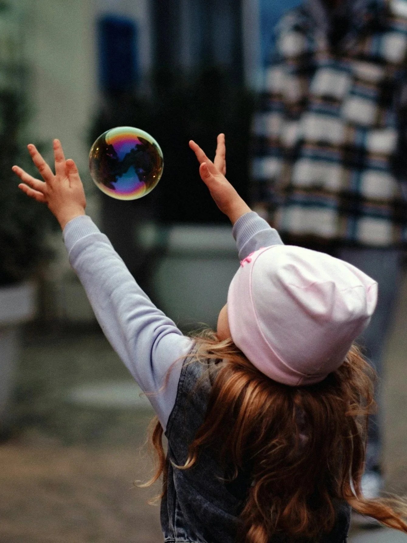 Ein Kind mit rotem Lockenhaar, das einen weißen Hut und eine graue Jacke trägt, greift nach einer schillernden Seifenblase in einer urbanen Umgebung.