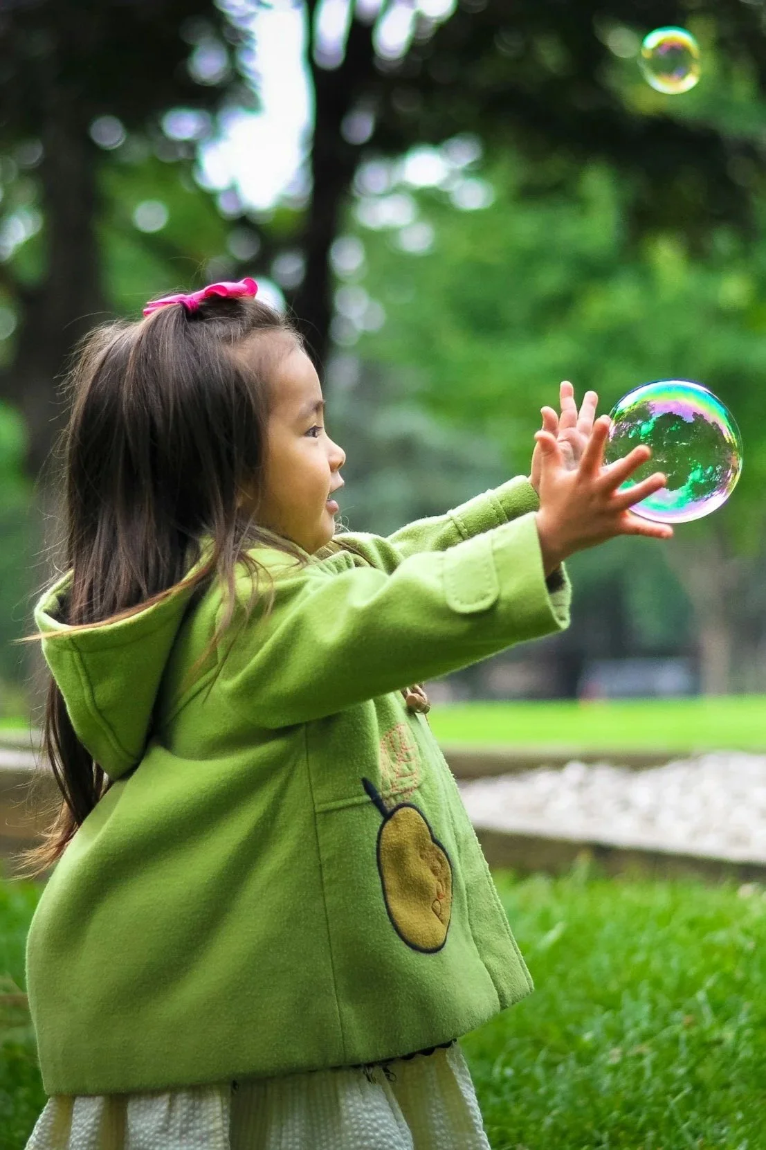 Ein Mädchen, das im Freien an einer Parkbank in einem grünen Park spielt, eine Seifenblase fängt und in die Luft schaut.