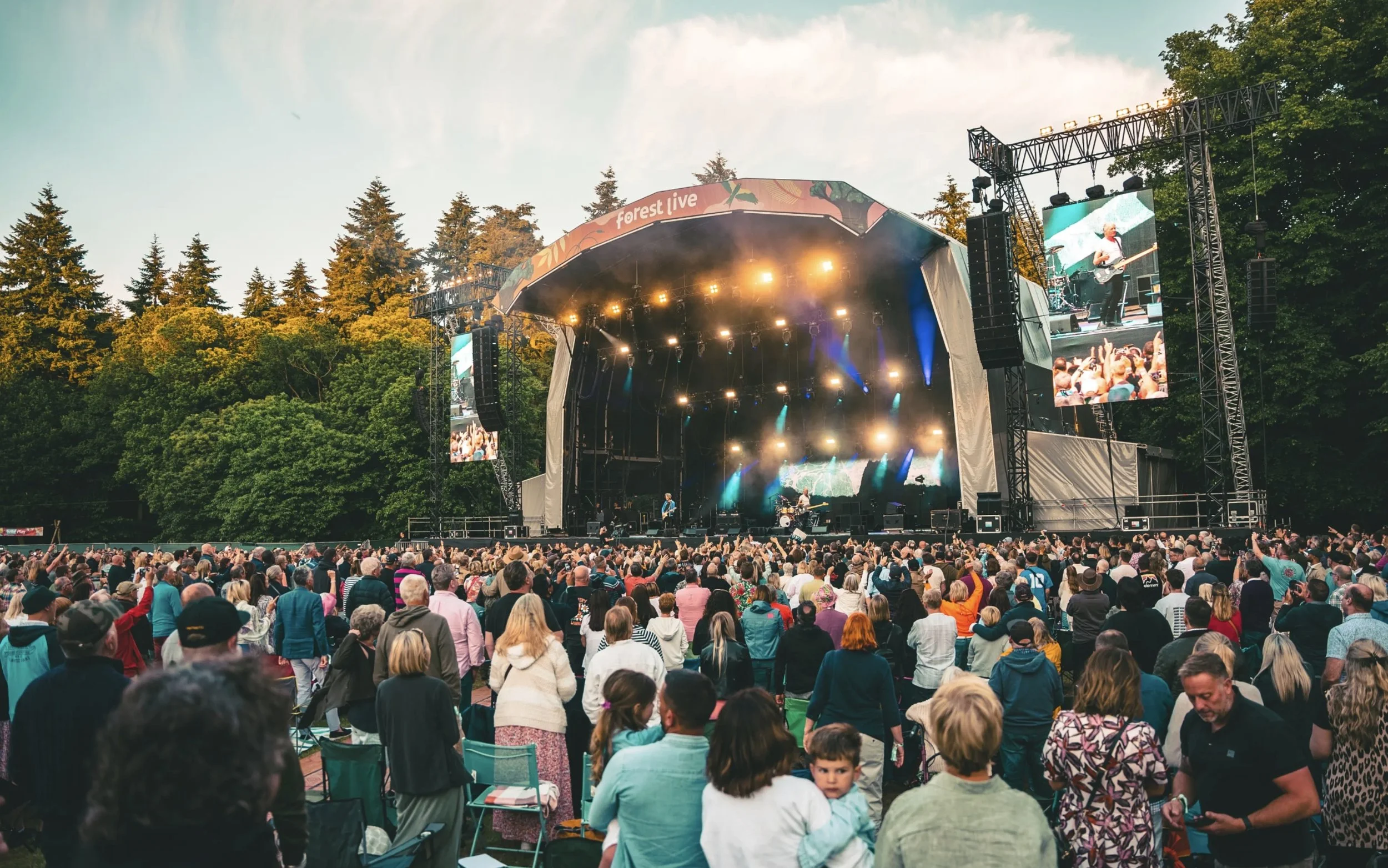 Families and fans gathered in front of the Forest Live stage on a summer evening, surrounded by tall trees.