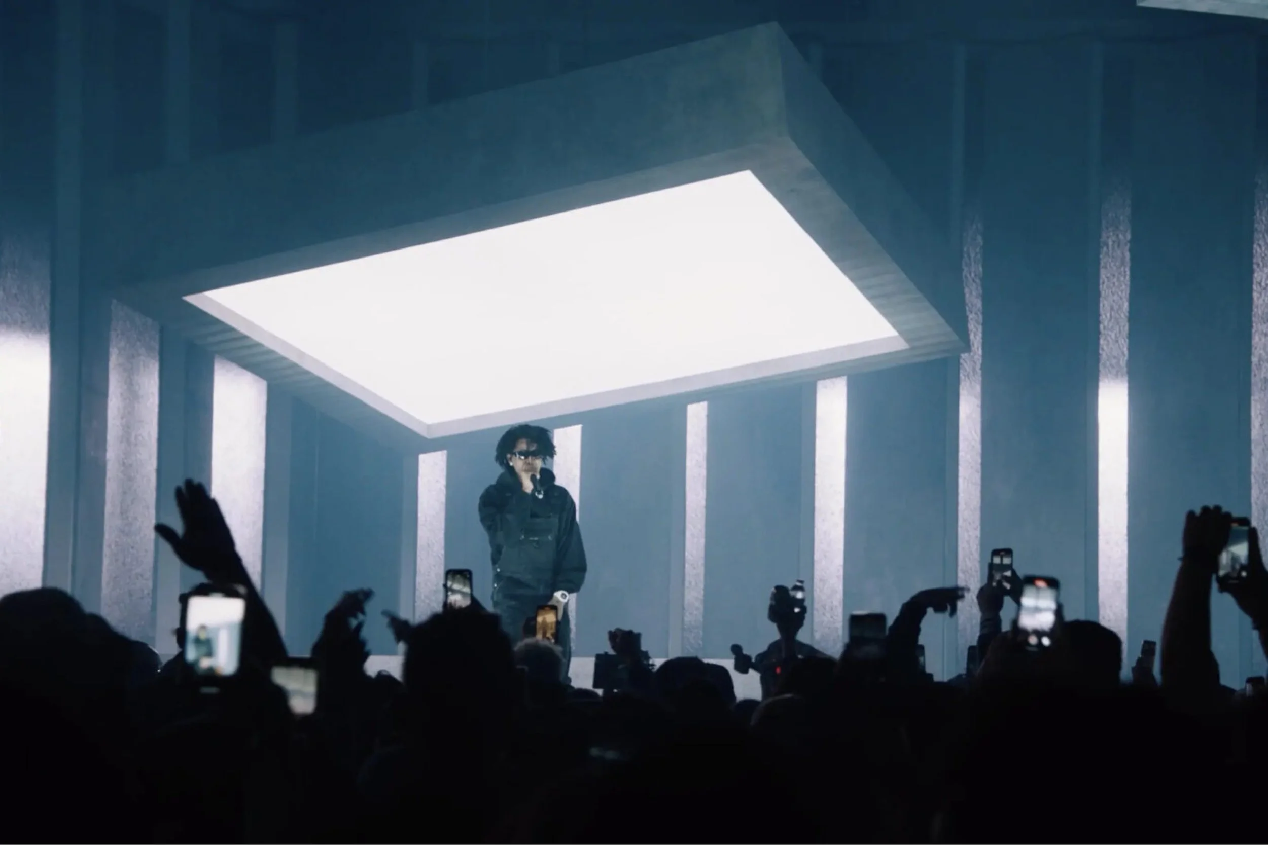 A performer on stage beneath a glowing light box as the crowd holds up their phones, from Cate Carter’s portfolio.