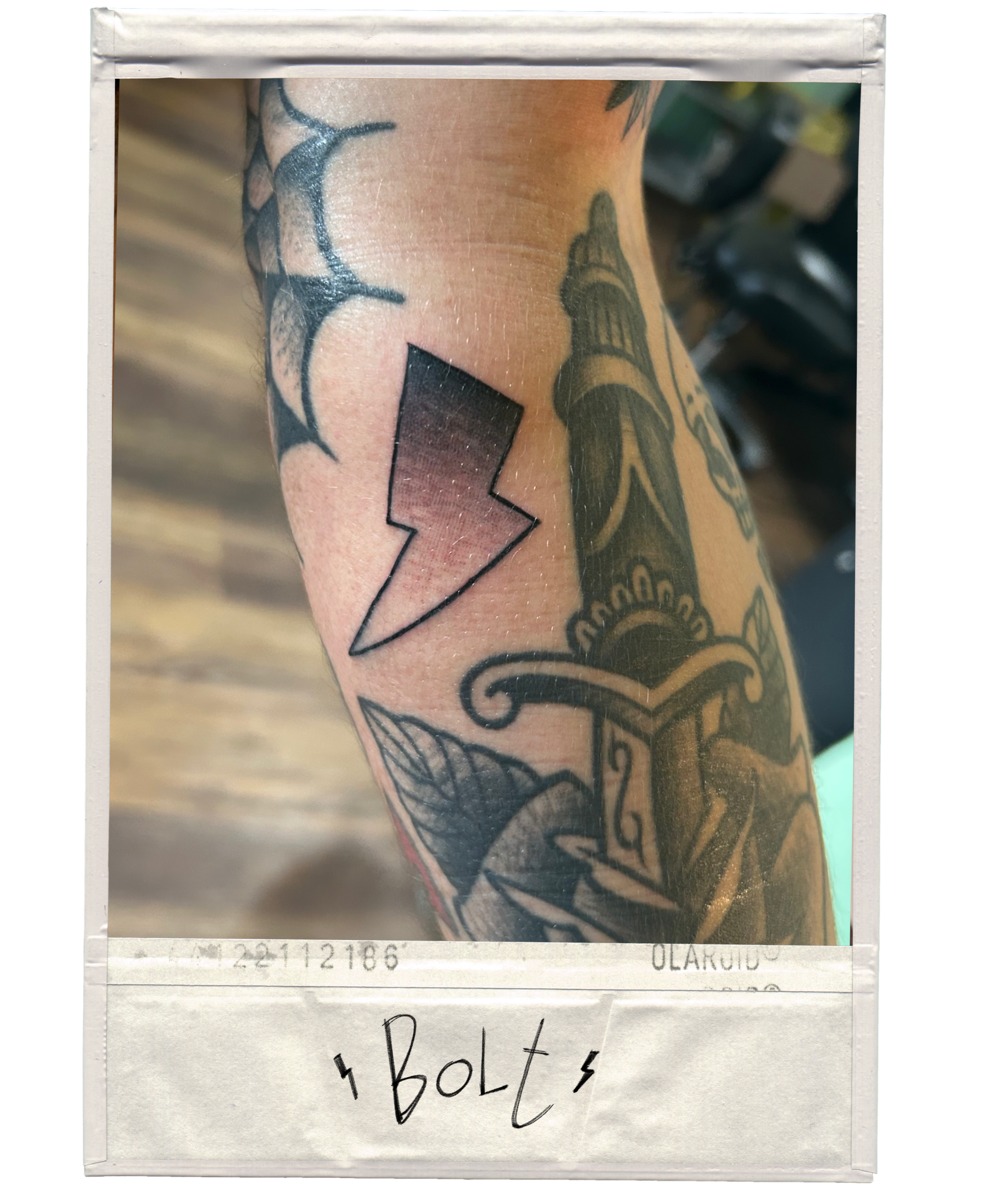 Tattooed arm with a lightning bolt design, a lighthouse, and decorative elements, with a Polaroid-style frame and the word 'Bolt' written at the bottom.