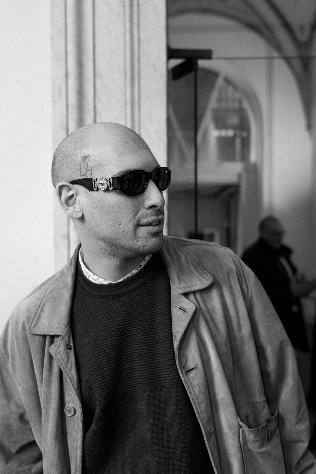 A man with a shaved head wearing dark sunglasses, a patterned shirt, and a jacket, standing indoors near a wall, with a blurred person in the background.