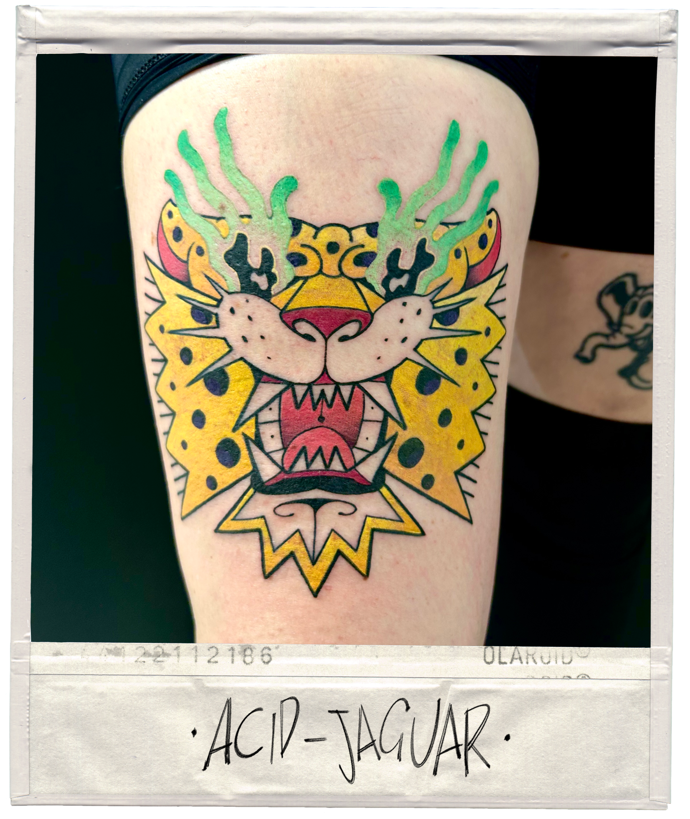 Tattoo of a colorful, stylized jaguar face with green flames, yellow spots, and an open mouth, on a person's thigh.