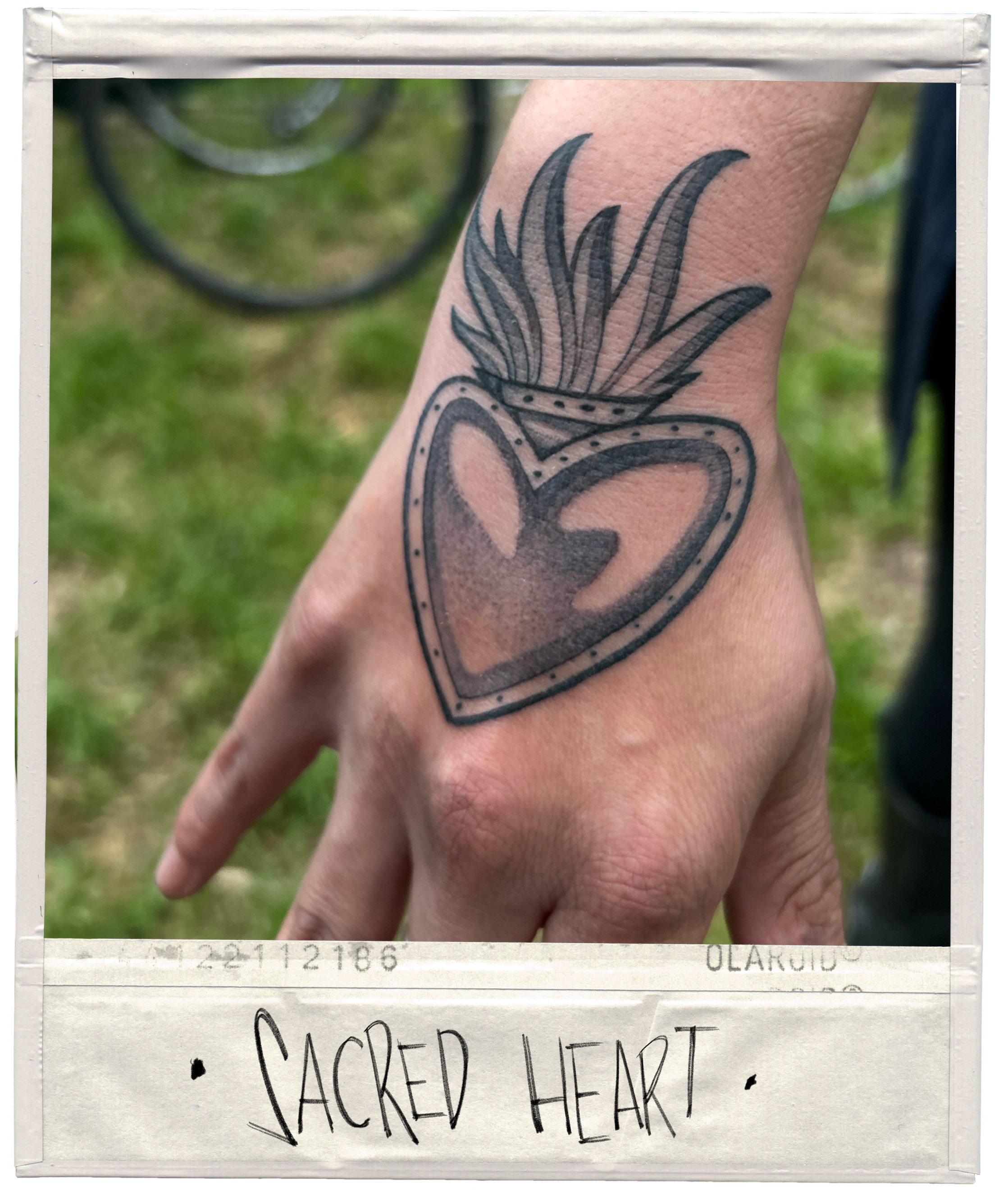 Tattoo of a heart with a flame or fire coming out of it on a person's hand, labeled 'Sacred Heart' on a note.